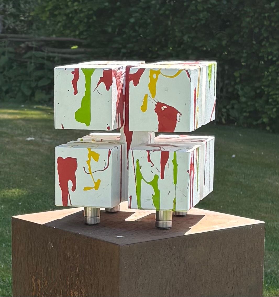Abstracte witte kubussculptuur met rode, groene en gele verfspatten op een houten sokkel in de buitenlucht.
