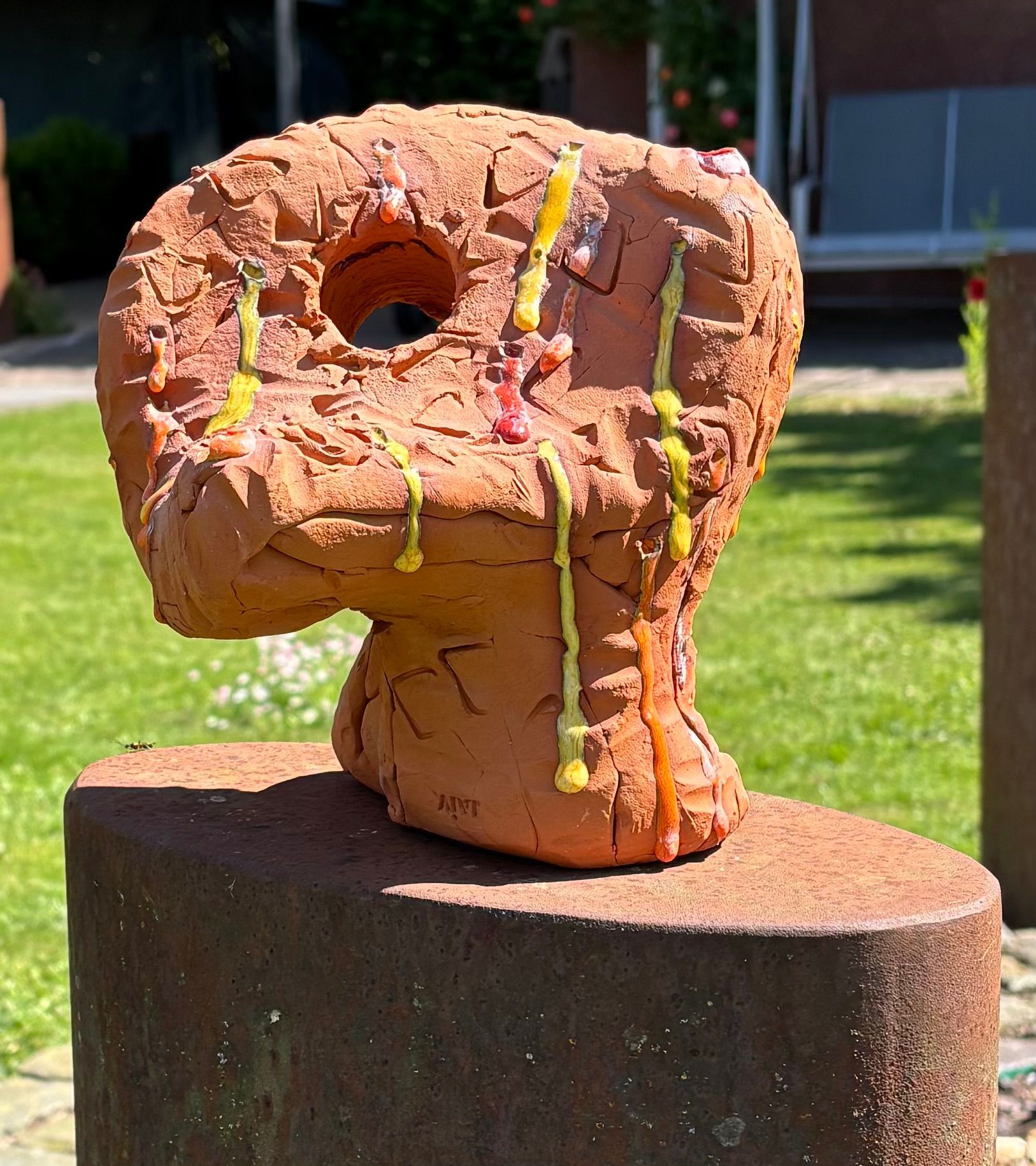 Roestkleurige abstracte sculptuur op een sokkel met gele verfspatten in een zonnige buitenomgeving.