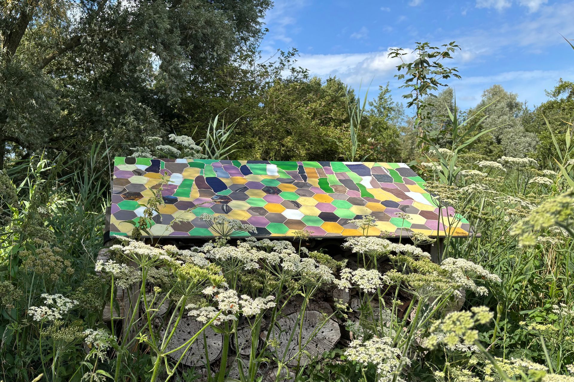 Kleurrijk schuurtje met mozaïekdak in een bloementuin onder een helderblauwe hemel.