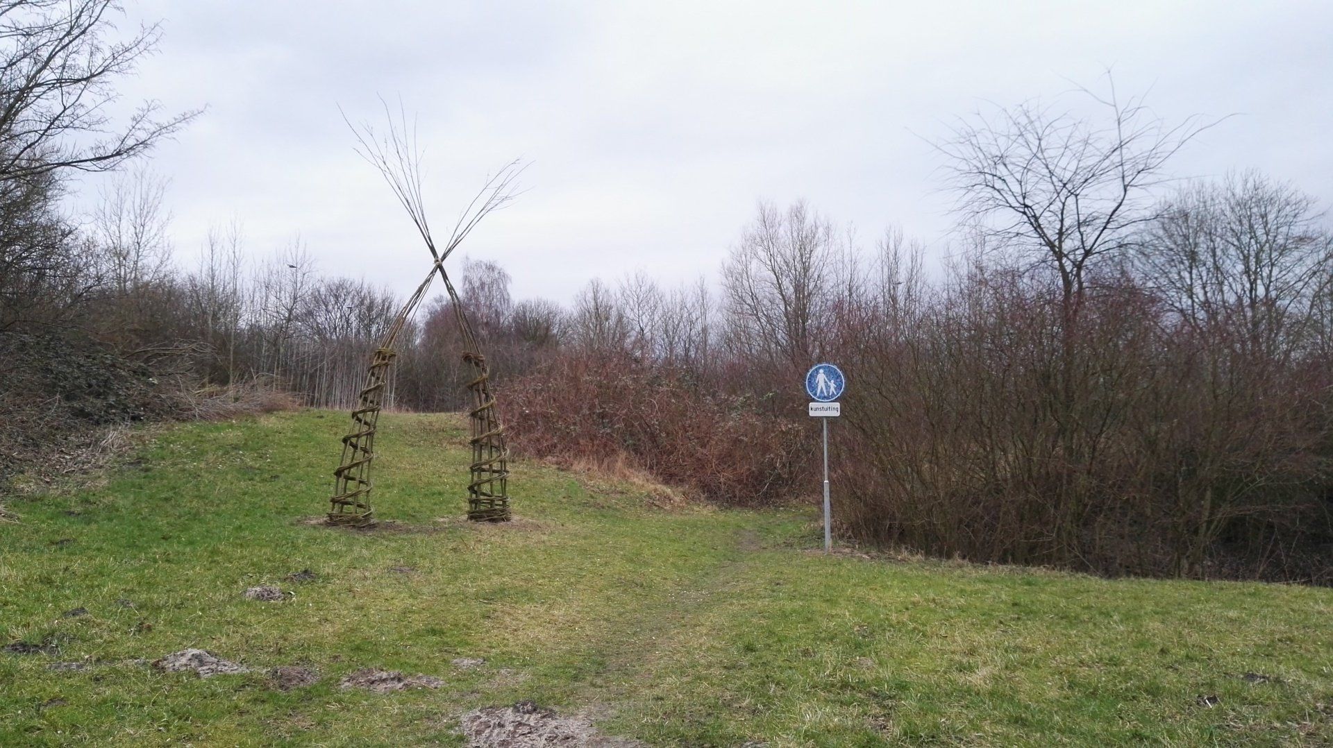 Een met gras begroeide heuvel met twee gebeeldhouwde houten palen en een bord naast kale bomen onder een bewolkte hemel.