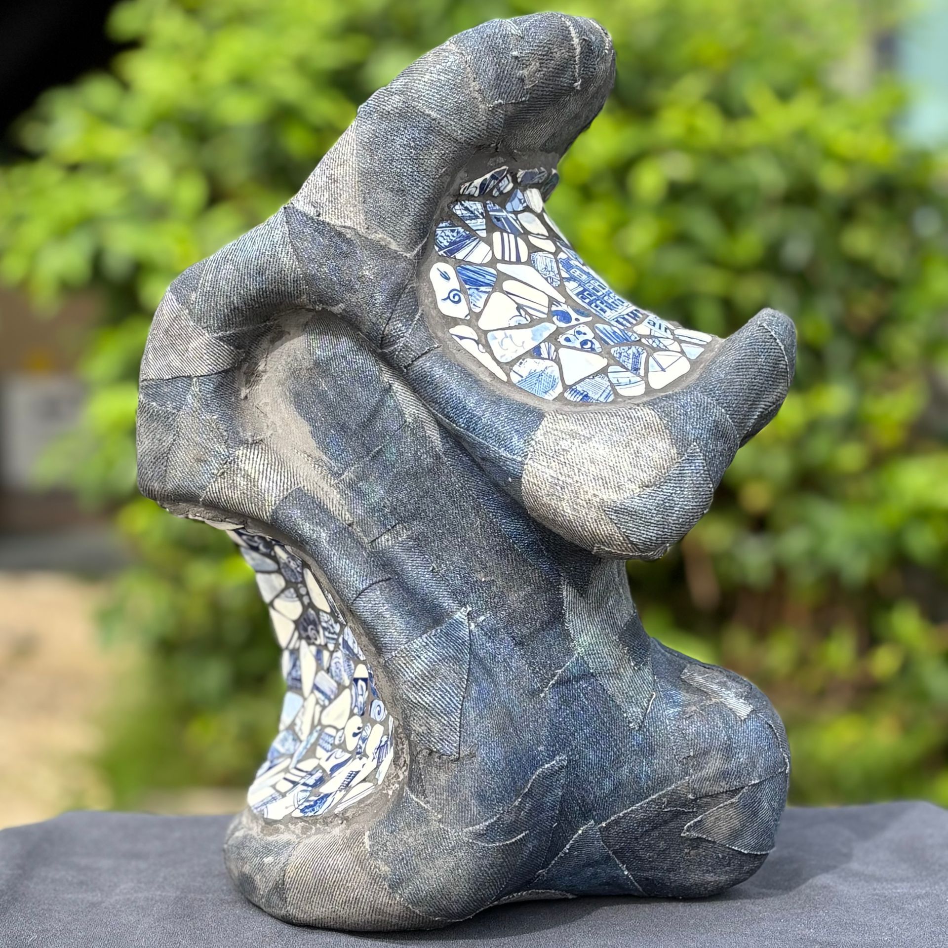 Abstracte blauw-grijze keramische sculptuur met een gestructureerd oppervlak, buiten tegen een wazige groene achtergrond.