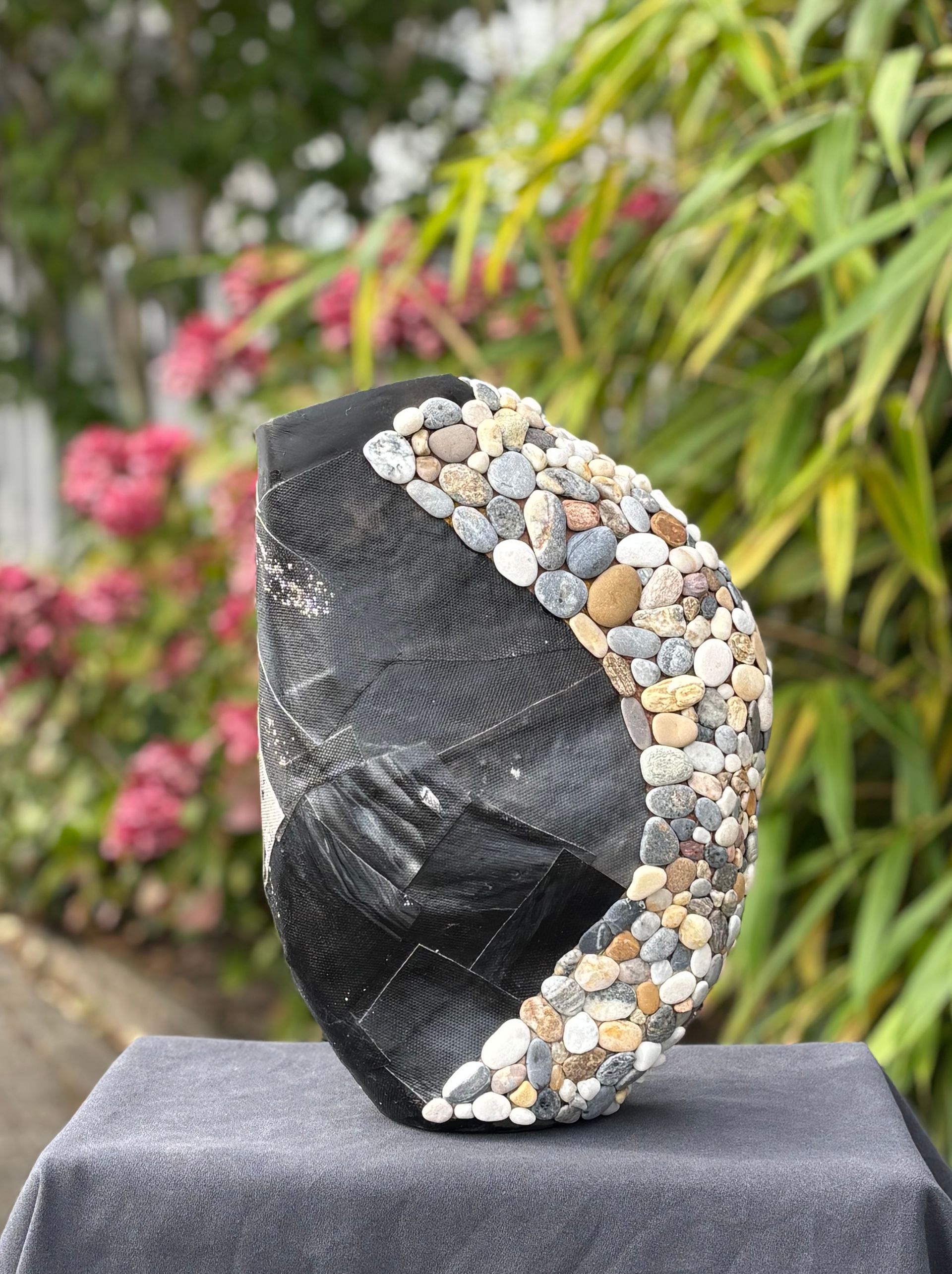 Zwarte, gestructureerde sculptuur met een gebogen rand, gevuld met kiezels, staat buiten op een sokkel.