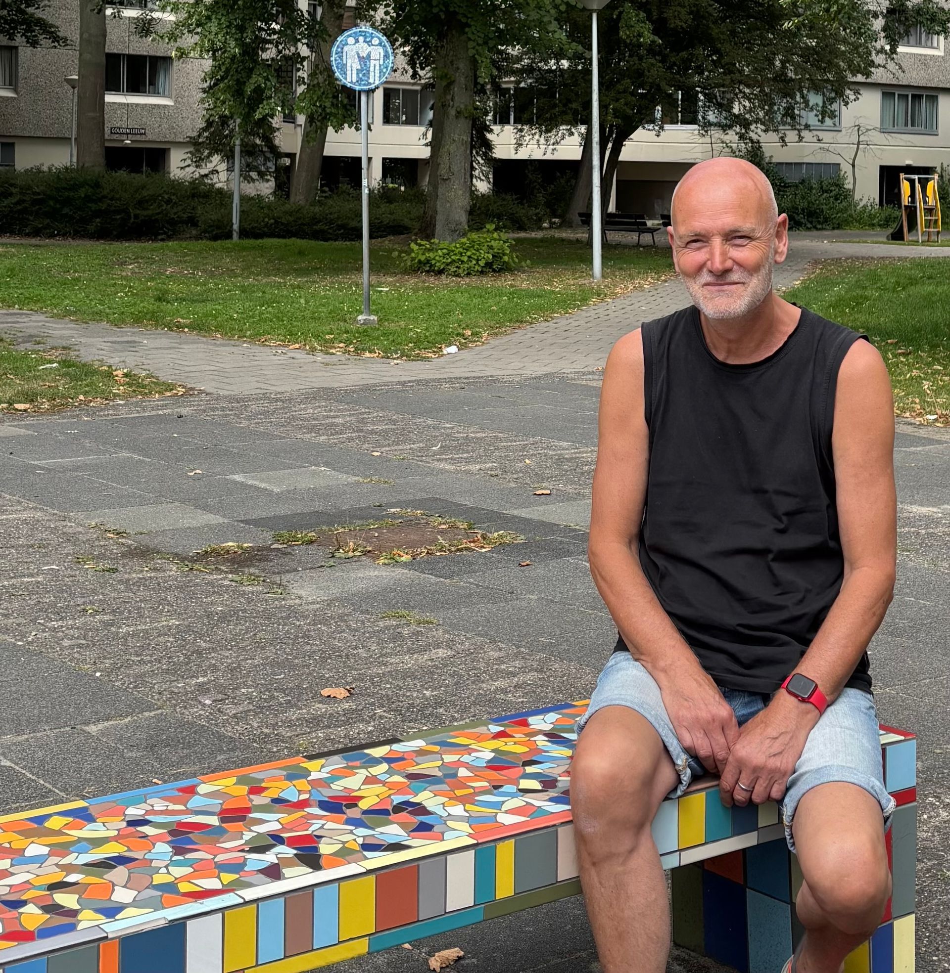 Een man zit op een kleurrijke mozaïekbank in een park, gekleed in een zwart mouwloos shirt en een spijkerbroekje.