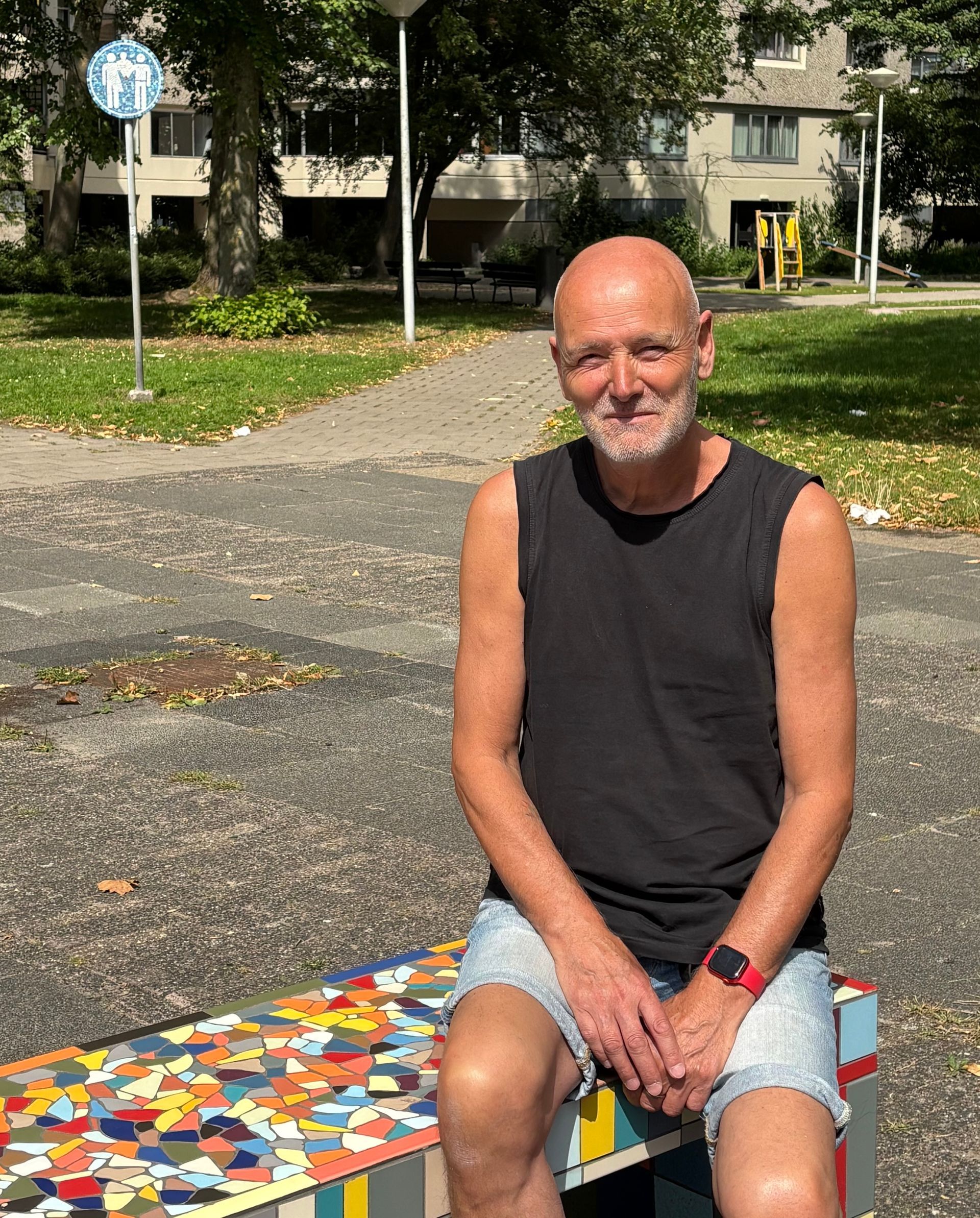 Een man in een zwart mouwloos shirt zit op een kleurrijke bank in een park en lacht naar de camera.