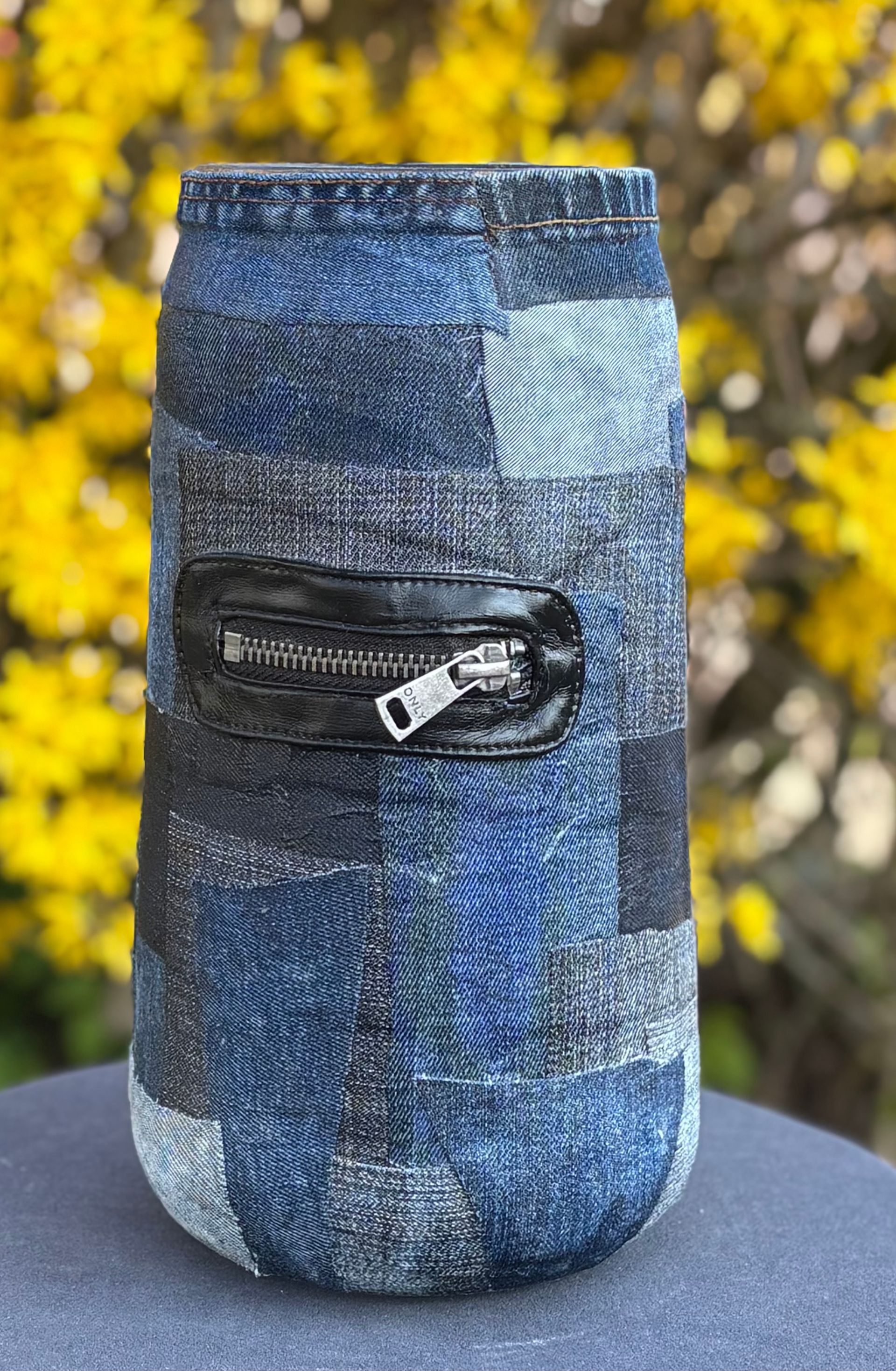 Blauwe patchwork denim tas met een zwart ritsvak, staand buiten tegen een wazige gele achtergrond.