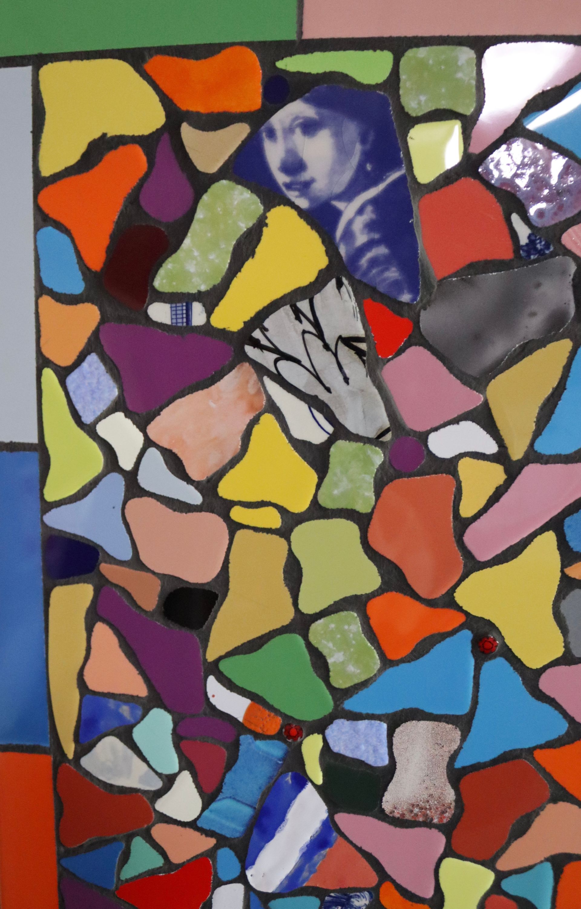 Glasmozaïek van kleurrijke abstracte stukken met een blauwgetint gezicht in het midden bovenaan.