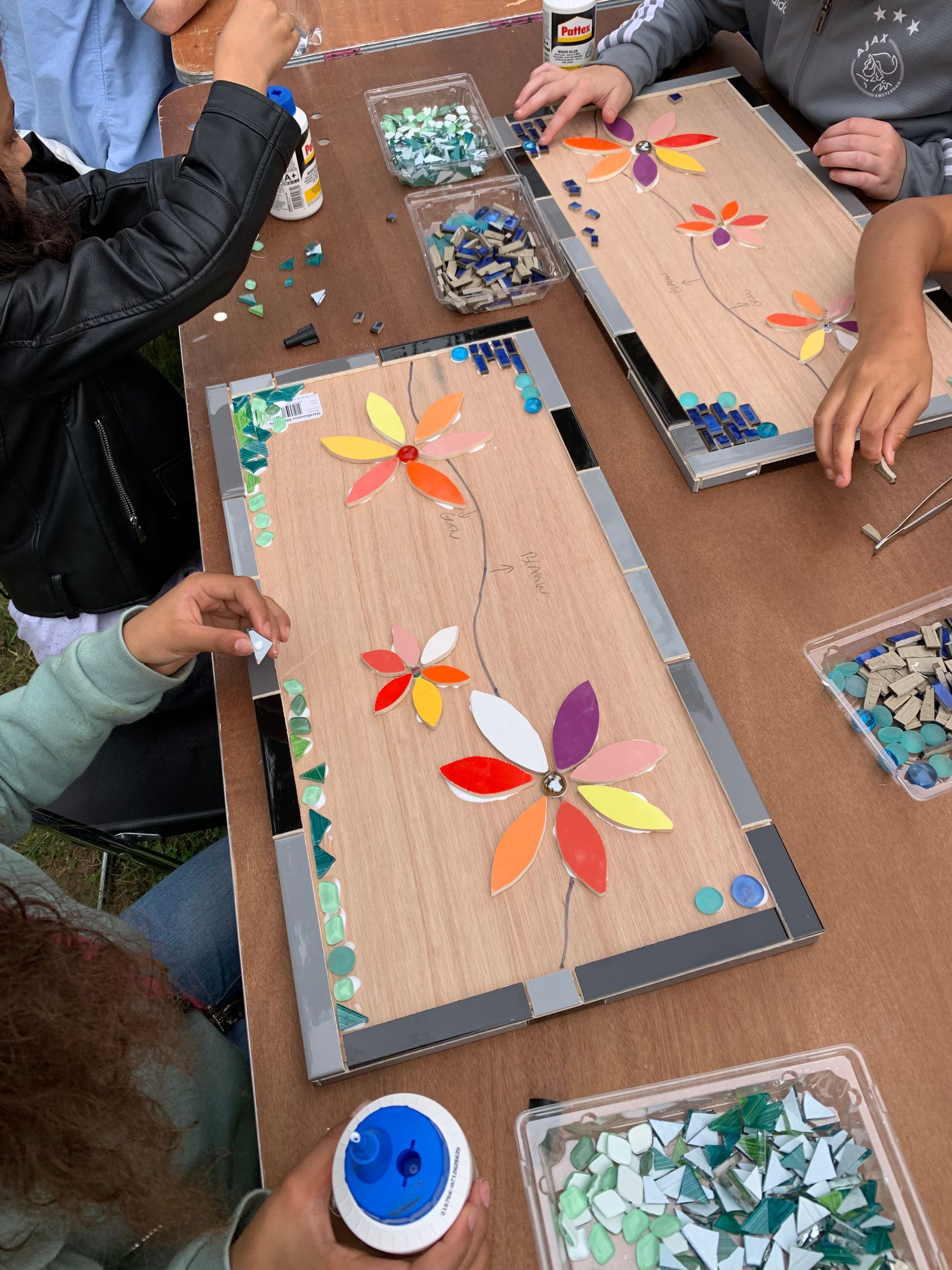 Mensen maken kleurrijke bloemenmozaïeken op kartonnen borden aan een knutseltafel.