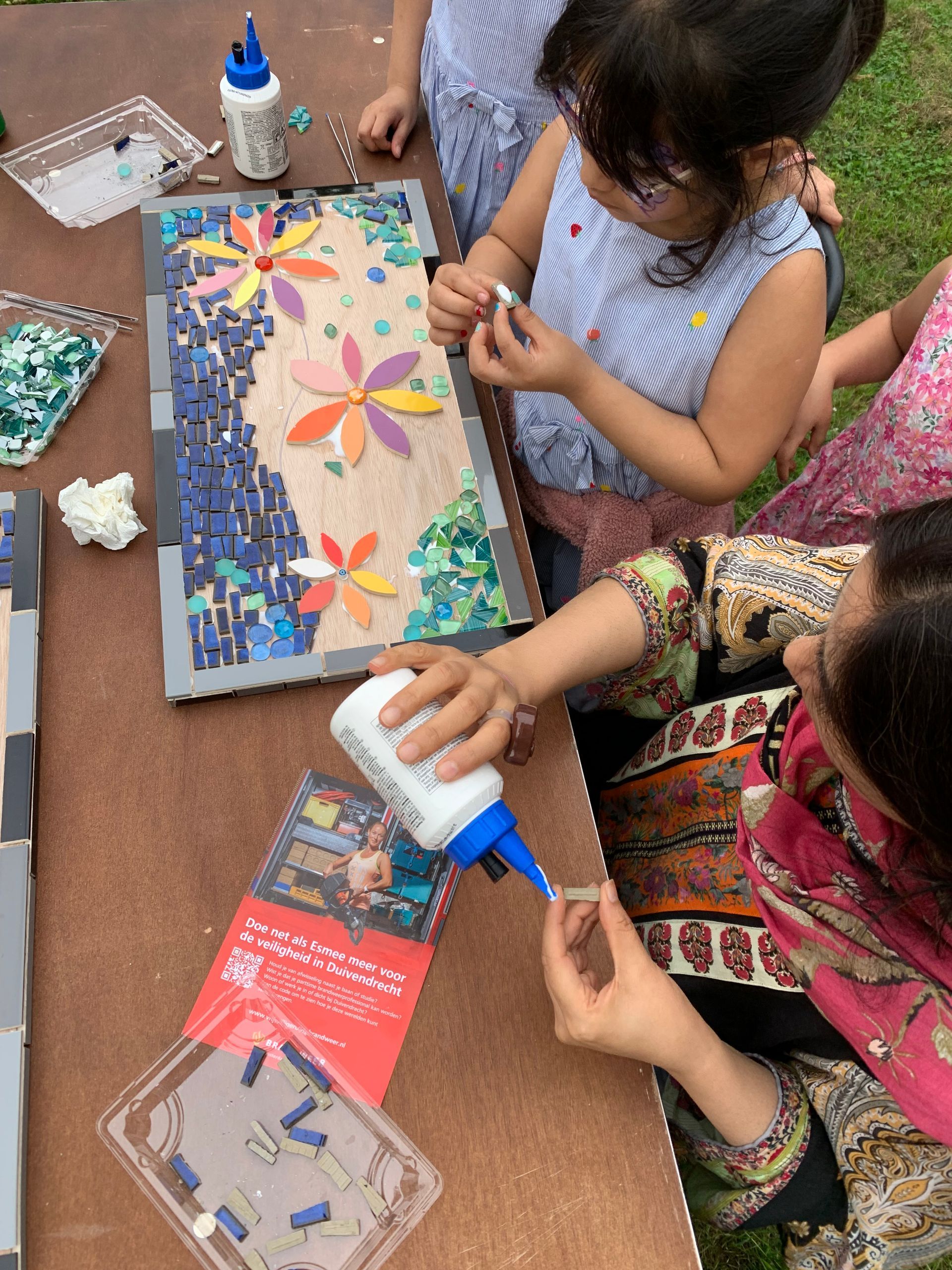 Kinderen maken kleurrijke stippenkunst op karton aan een knutseltafel met lijmflesjes en geverfde patronen.