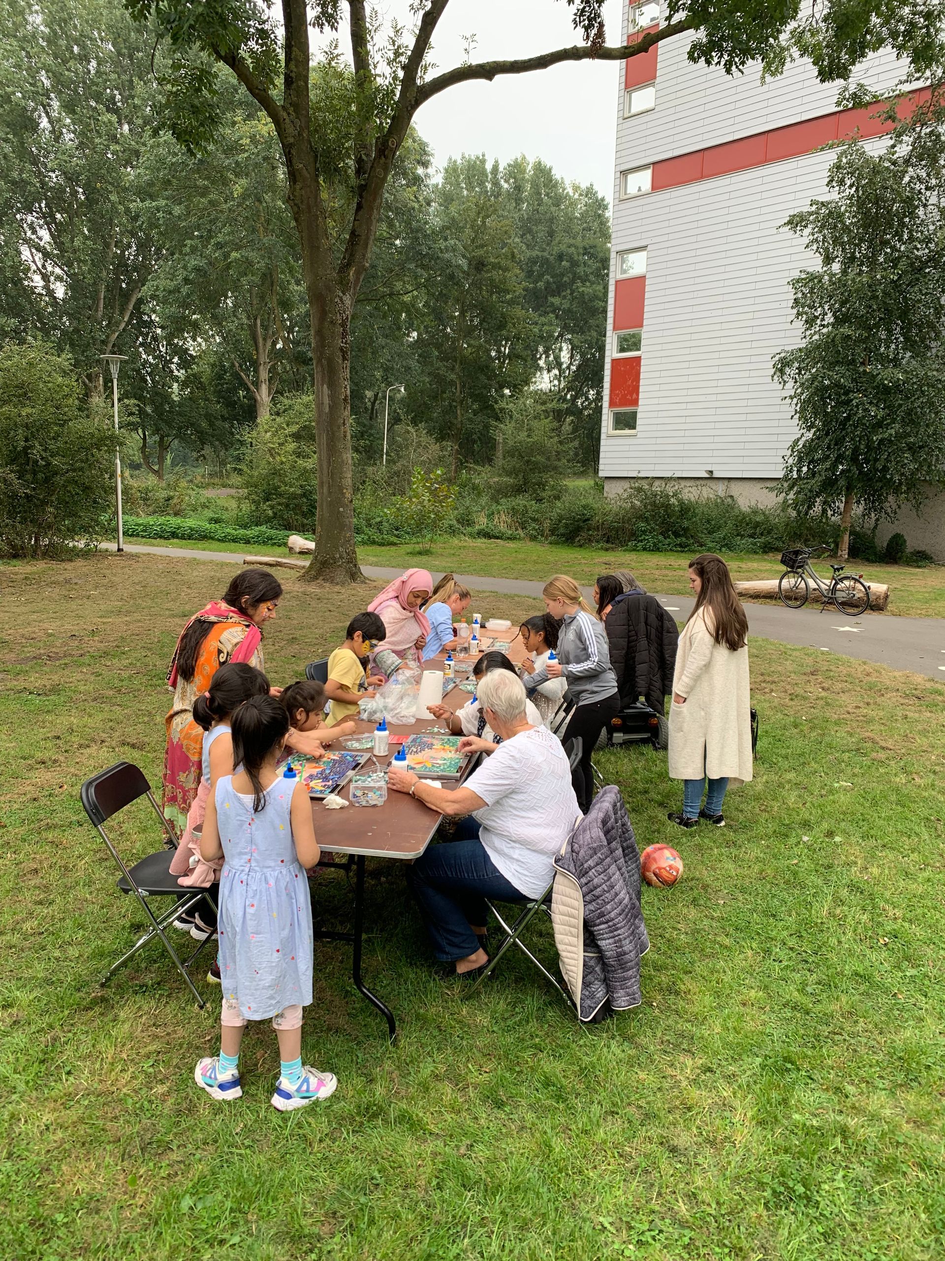 Kinderen en volwassenen verzamelden zich rond een picknicktafel in een grasrijk park, waar ze samen aten en dronken.
