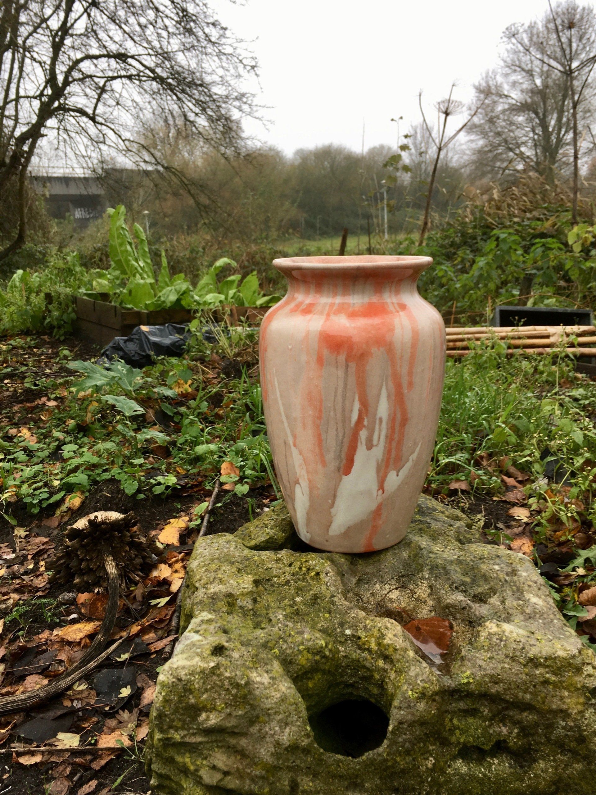 Grote roze-witte keramische vaas op een steen in een tuin, met bomen en groen op de achtergrond.