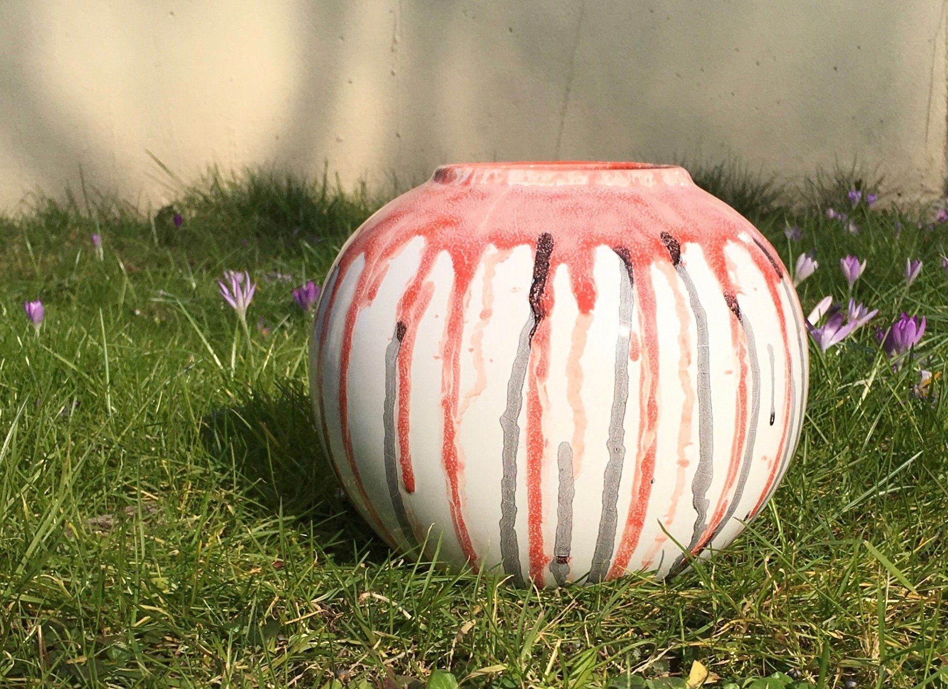Witte ronde plantenbak met rode druppels, geplaatst op gras tegen een beige muur, omringd door paarse bloemen.