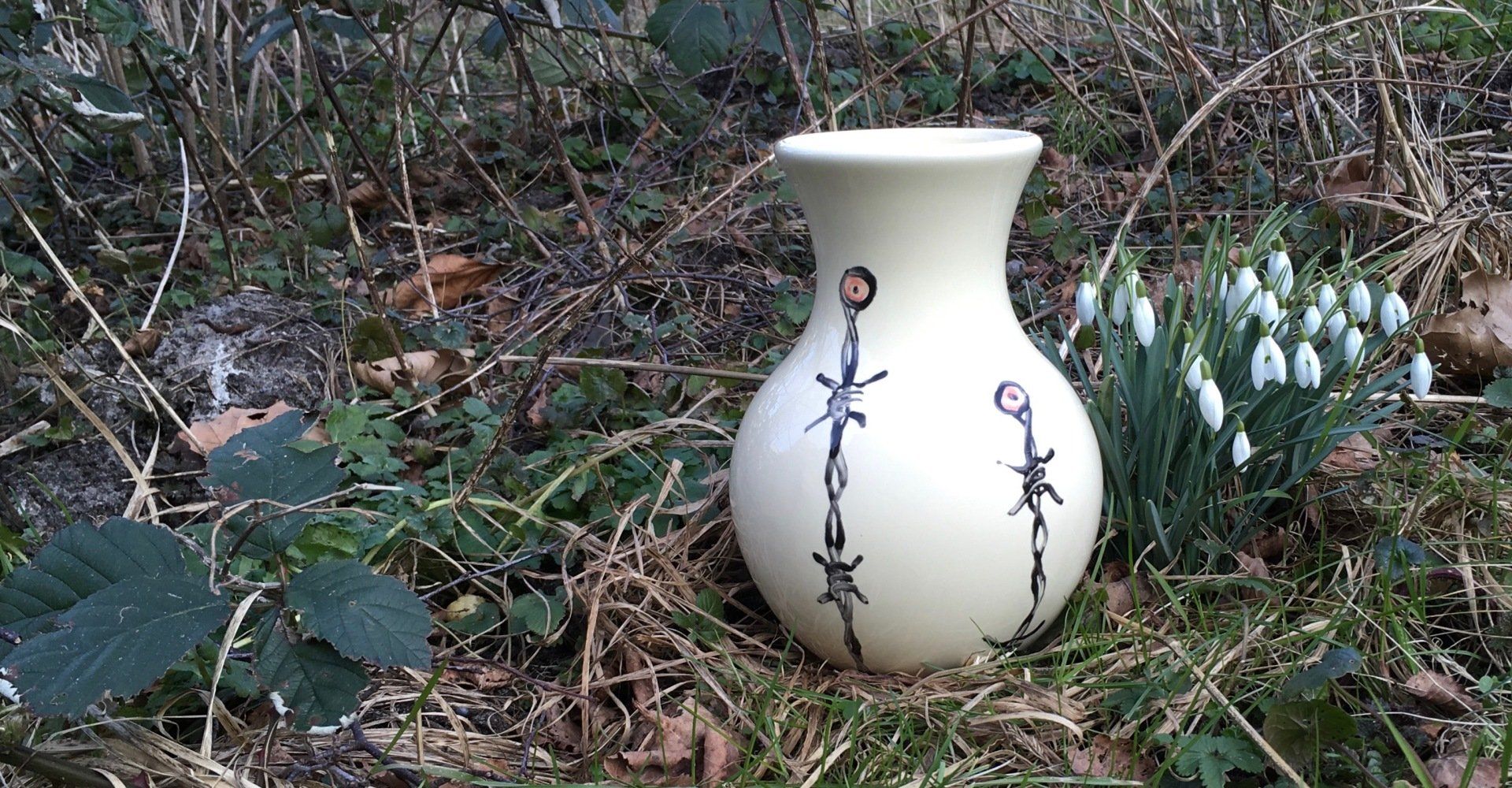 Witte keramische vaas met blauwe rankmotieven, te midden van tuinplanten en droge bladeren.