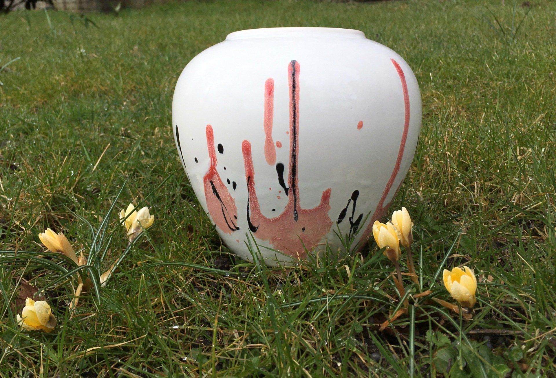 Witte vaas met rode druppels, staand in het gras tussen kleine gele bloemen.