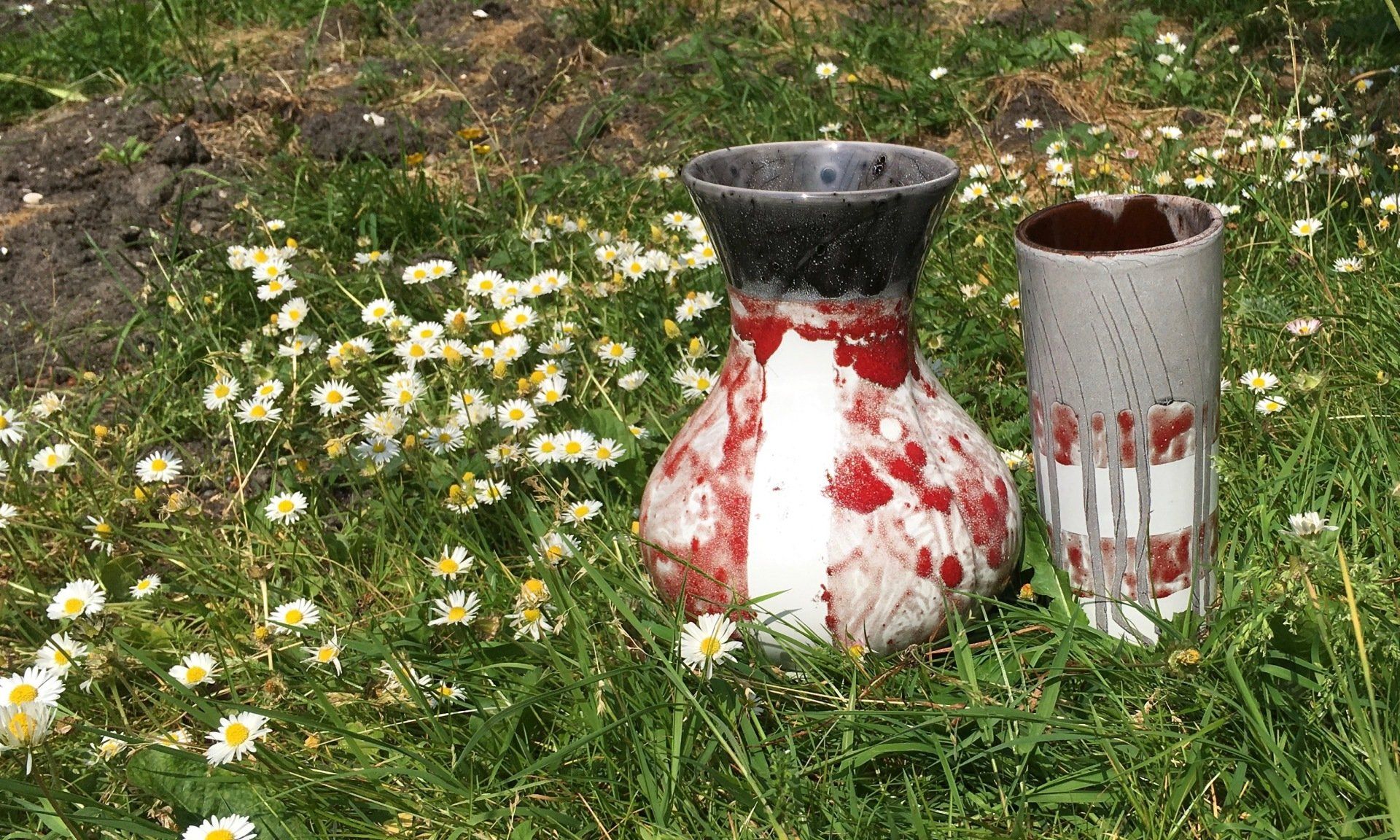 Twee verweerde rood-witte vazen ​​in een grasveld met wilde bloemen.