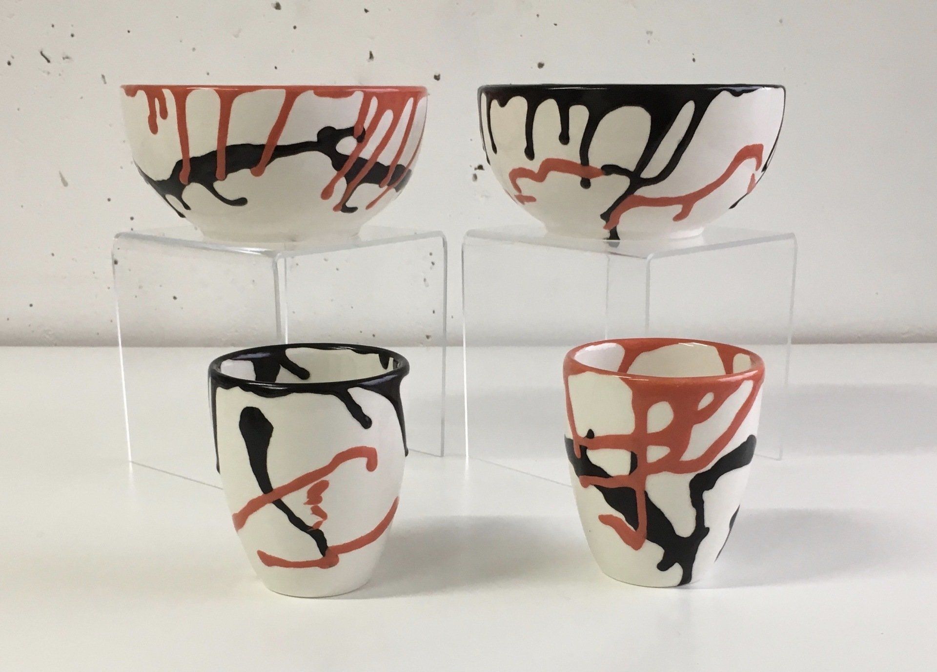 Vier abstracte, beschilderde glazen bekers op een witte achtergrond, met rode, zwarte en crèmekleurige druppels.