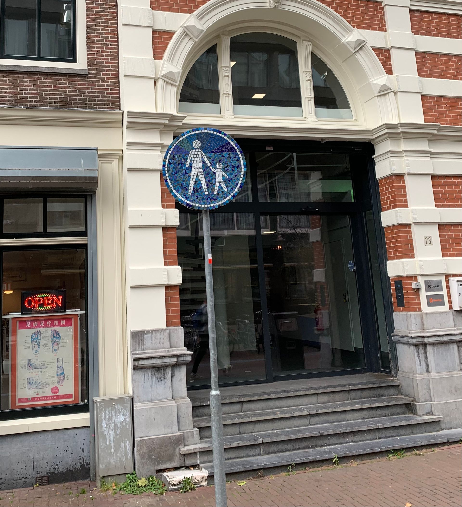 Ingang van een bakstenen gebouw met trappen en een blauw rond bord op een paal ervoor.