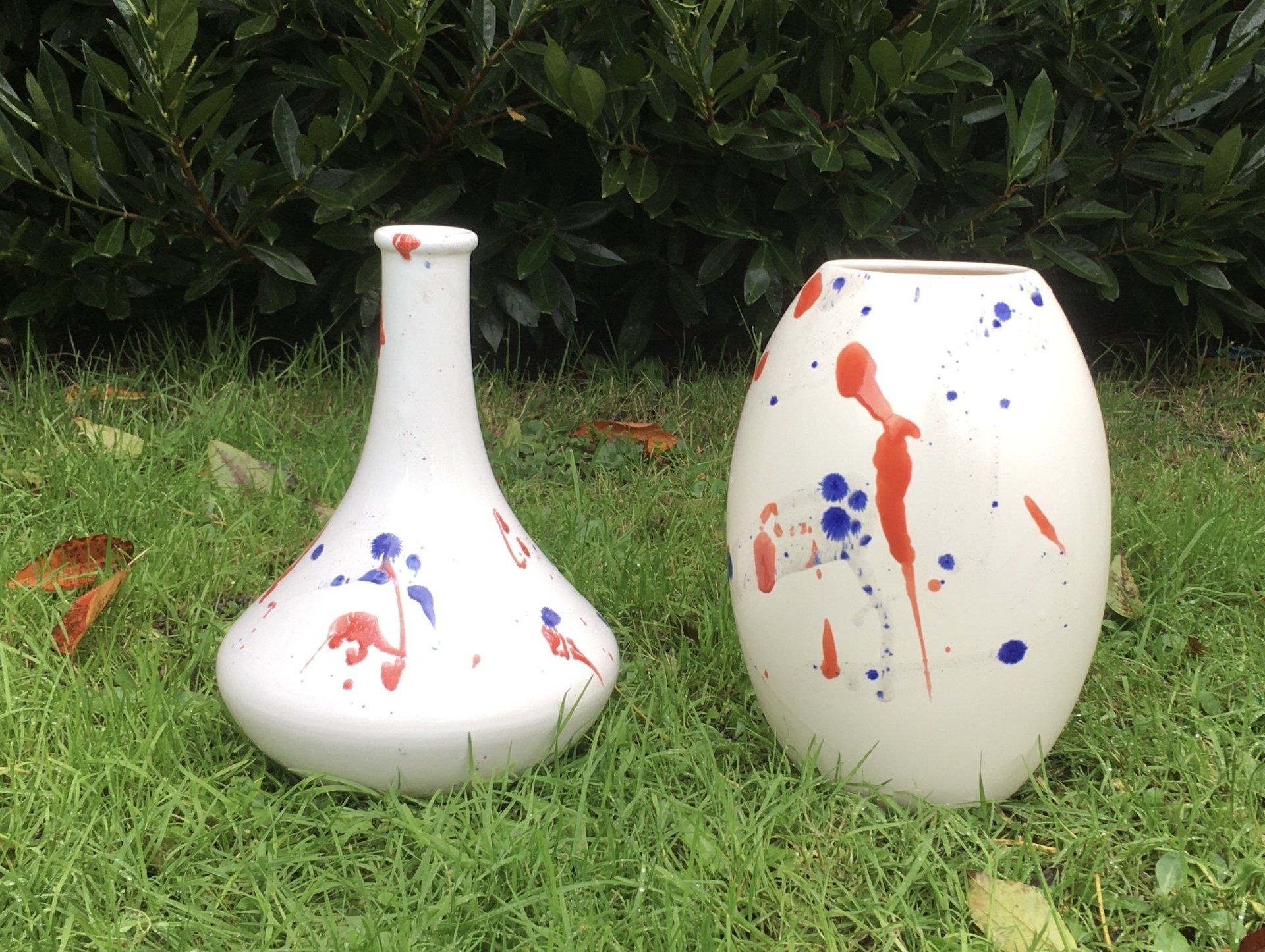 Twee witgeschilderde keramische vazen ​​met kleurrijke spetters op het gras voor struiken.