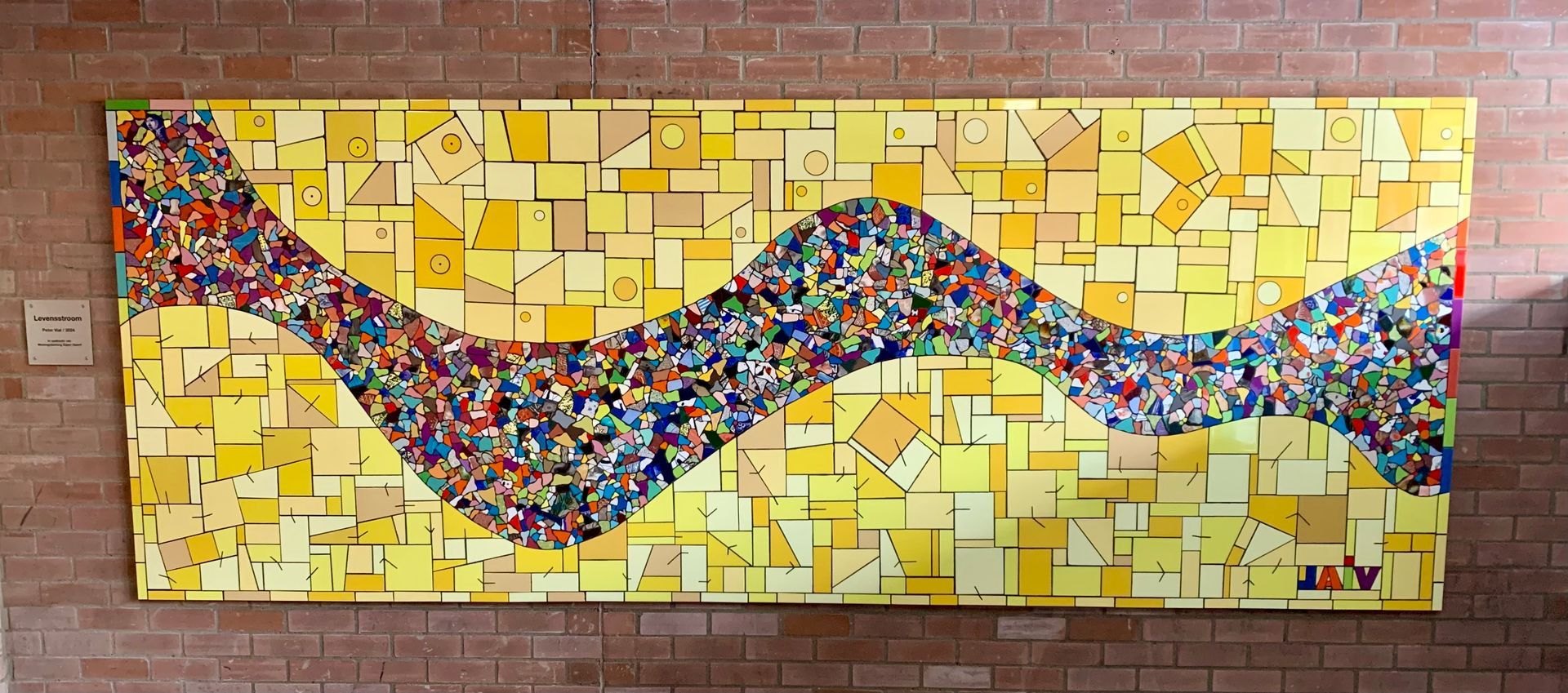 Abstracte mozaïekmuurschildering van kleurrijke mozaïekstukjes die een golvende lijn vormen op een gele bakstenen muur.