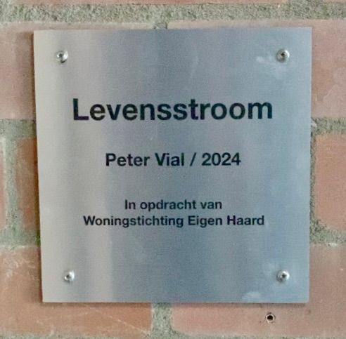 Silver plaque on brick wall reading “Levensstroom,” with “Peter Vial / 2024” and “Woningstichting Eigen Haard.”