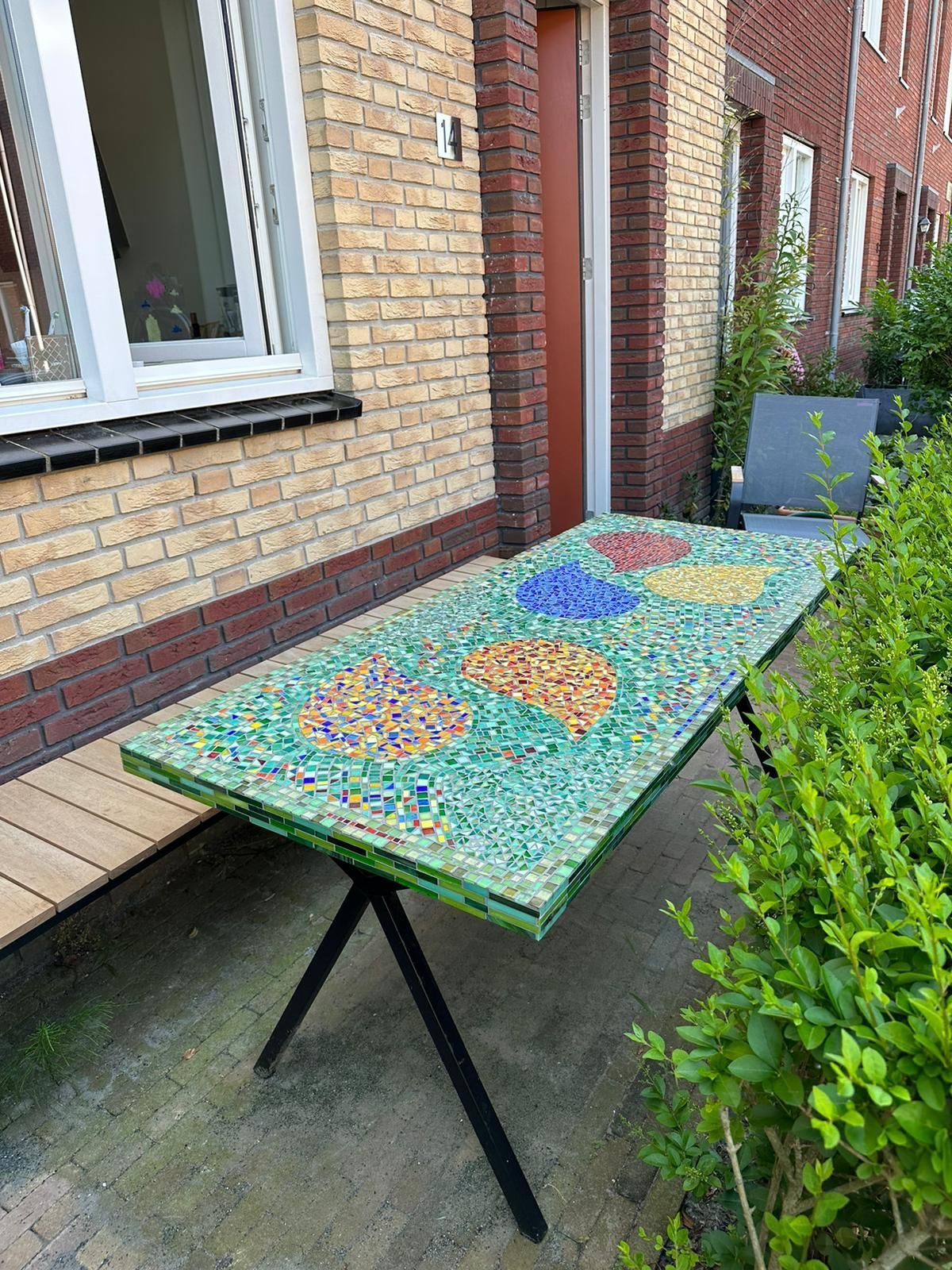 Tafel met mozaïekblad naast een bakstenen gebouw en een heg, met kleurrijke glaspatronen op een groene voet.