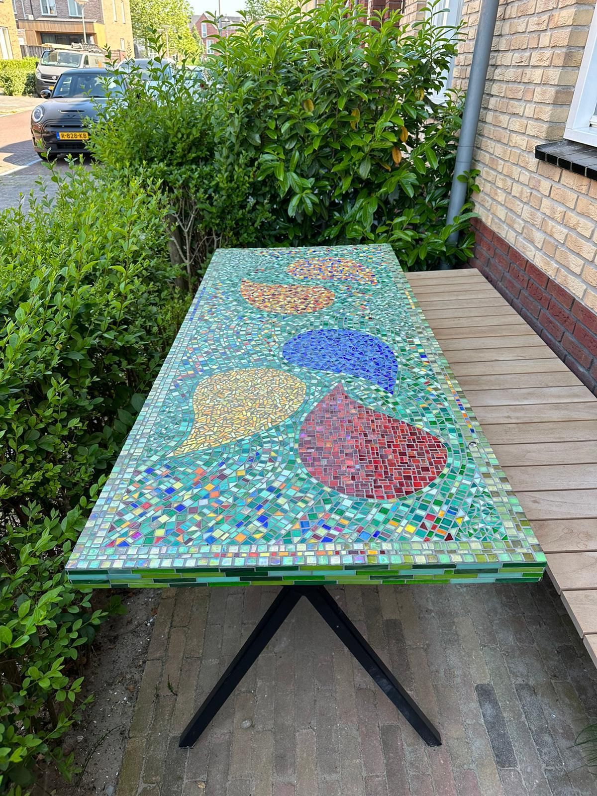 Een buitentafel met mozaïekblad op een terras naast een bakstenen muur en een heg.