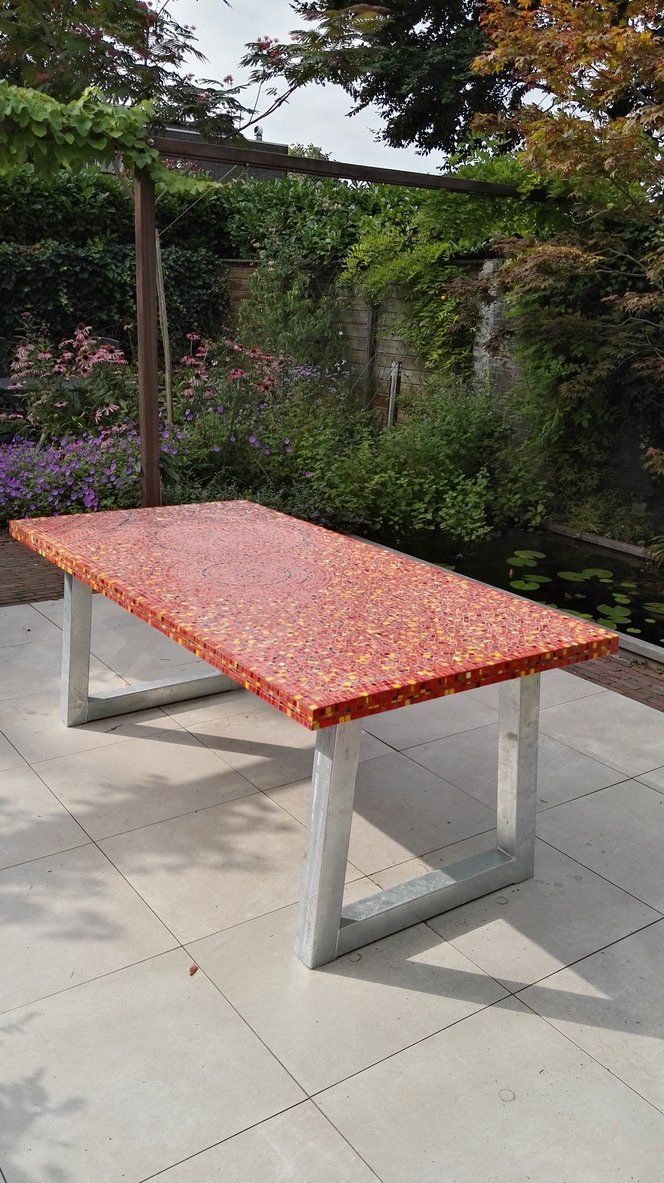 Buitentafel met een rood mozaïekblad en witte poten op een terras te midden van groen.