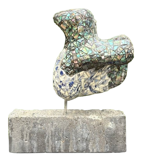 Abstracte mozaïeksculptuur op een betonnen sokkel, met een iriserend tegeloppervlak tegen een witte achtergrond.