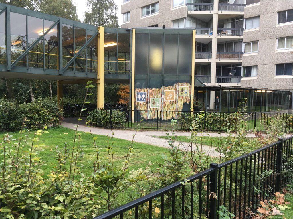 Binnenplaats met zwart hek, groene tuin, glazen loopbrug en appartementencomplexen op de achtergrond.