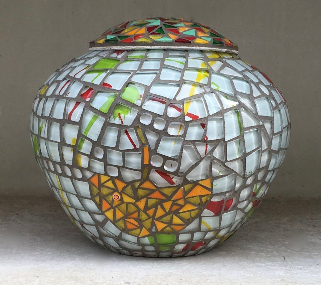 Mozaïek glazen vaas met kleurrijke driehoekige en witte mozaïeksteentjes op een grijze achtergrond.