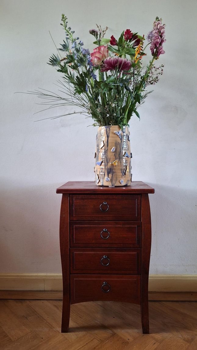 Een hoge vaas met gemengde bloemen staat op een klein houten kistje tegen een kale muur.