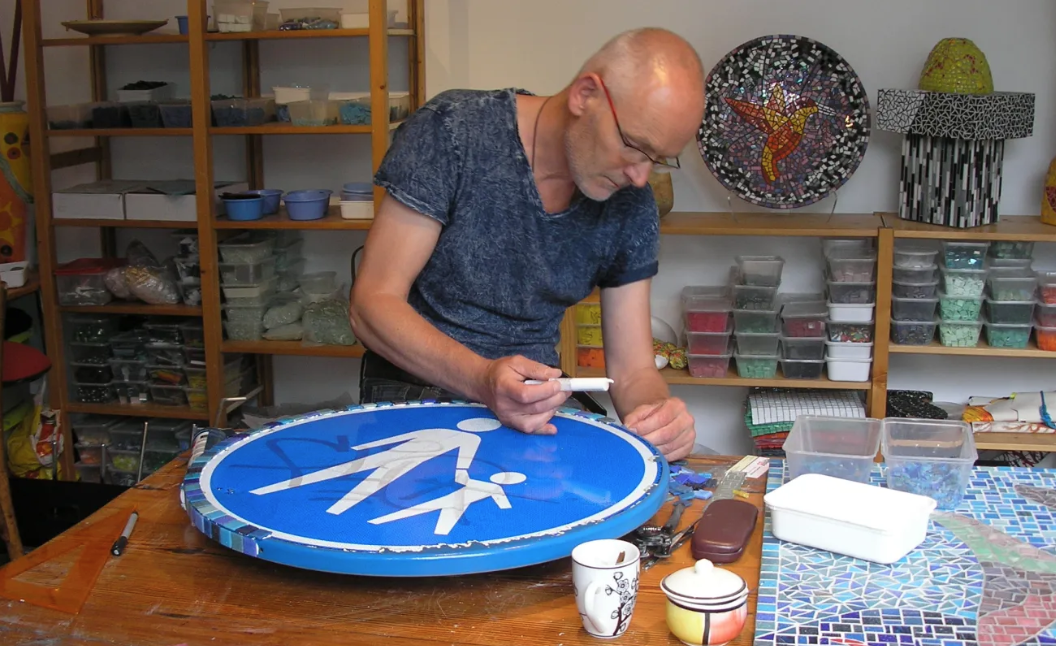 Een kunstenaar maakt mozaïek aan een werktafel in een atelier, met materialen en kleurrijke kunstwerken achter hem.