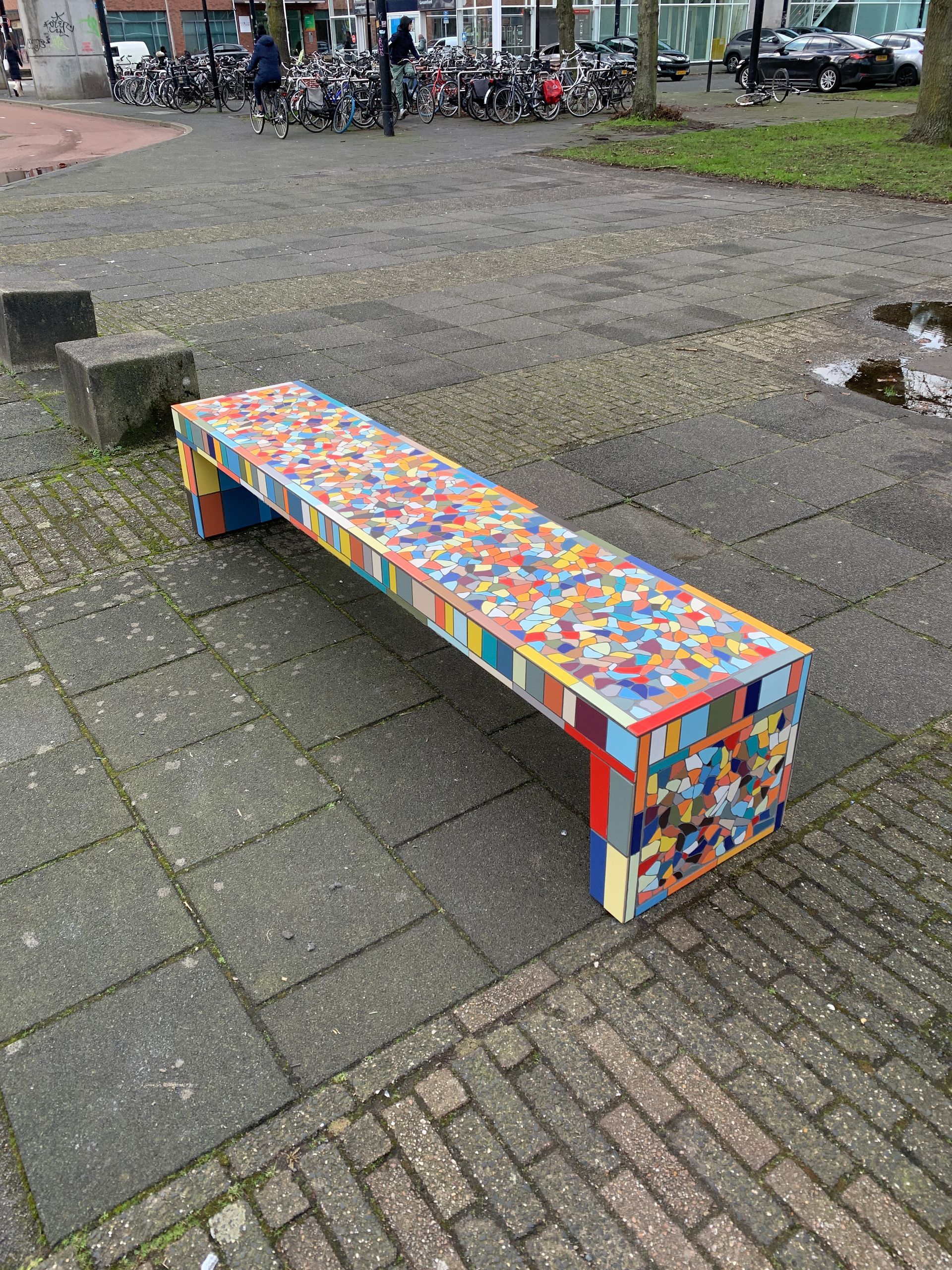 Een kleurrijke mozaïekbank op een geplaveid buitenplein, vlakbij fietsen en een laag stenen blok.
