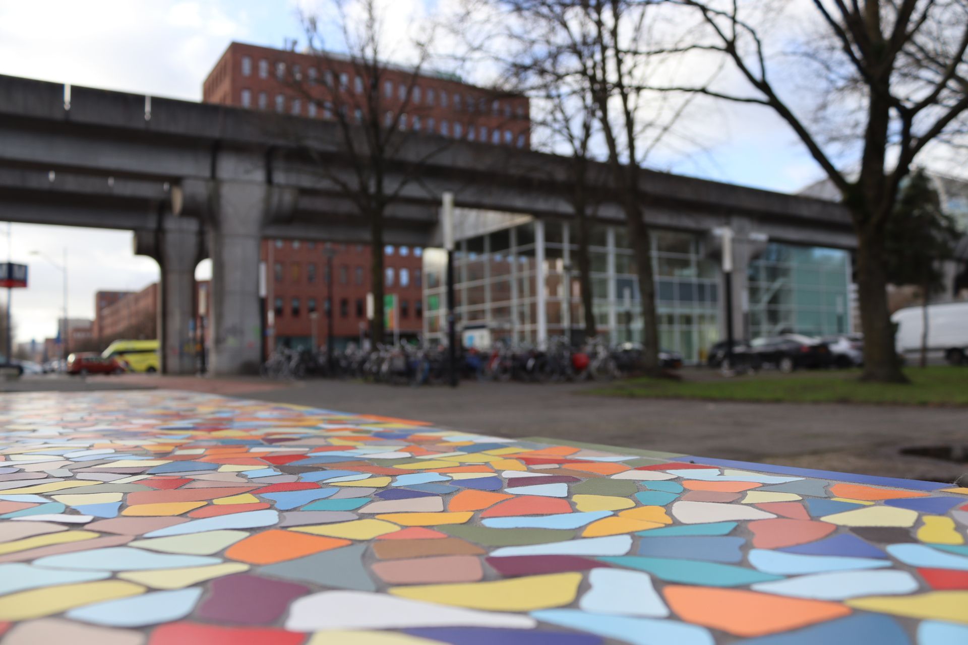 Kleurrijke mozaïekbestrating op de voorgrond, met daarachter een stadsgebouw en een verhoogd spoor.