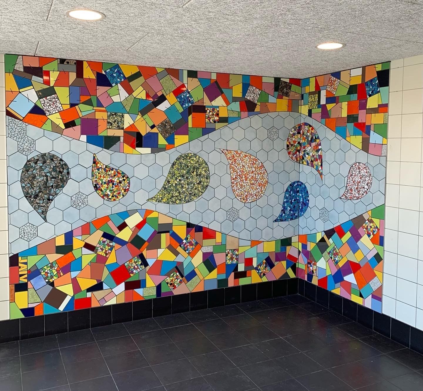 Een kleurrijk abstract mozaïekmuurschilderij op een hoekmuur met visachtige vormen en een witte tegelachtergrond.