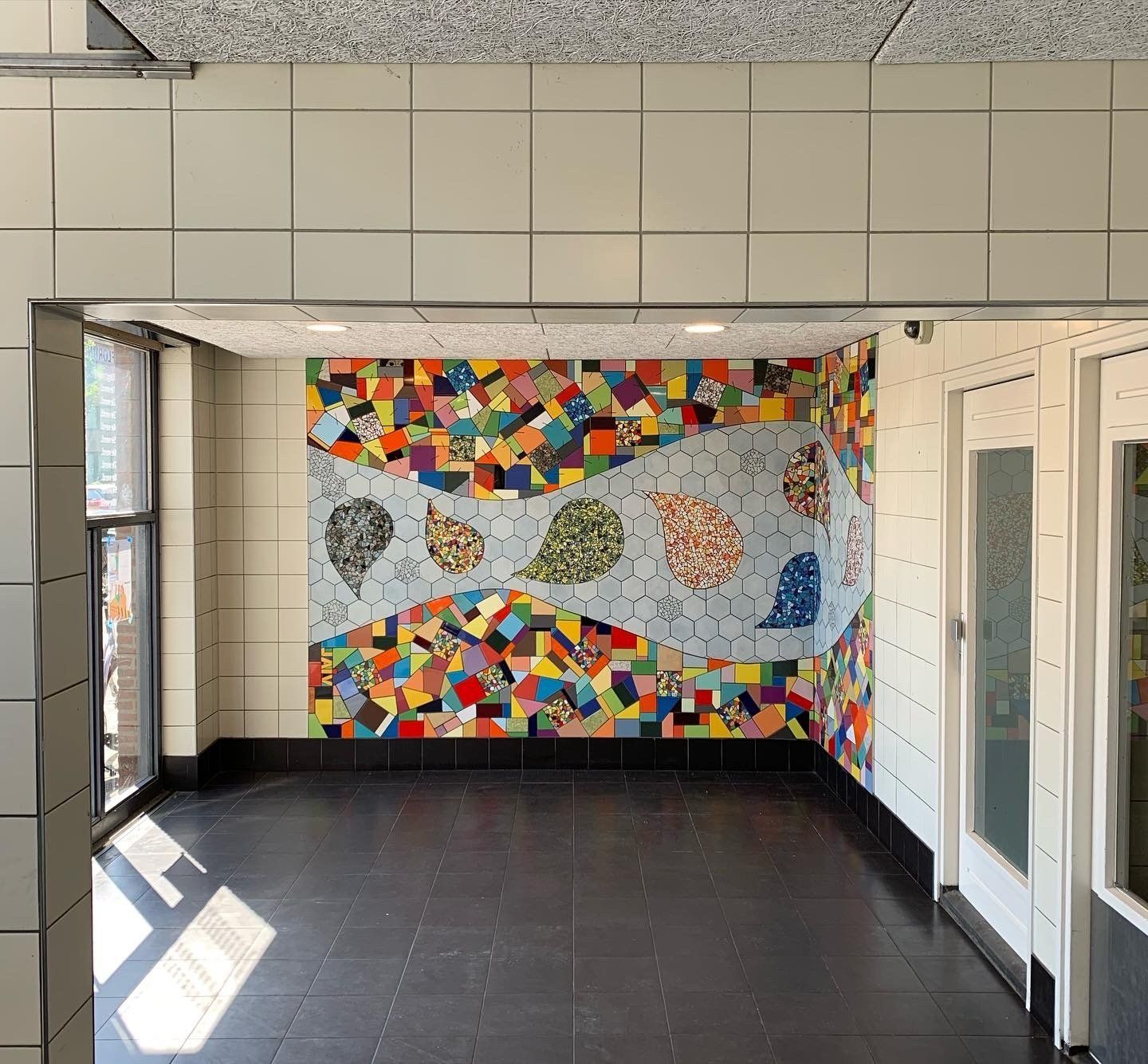 Gang met een kleurrijk mozaïek op de achterwand en een donkere tegelvloer onder fel zonlicht.
