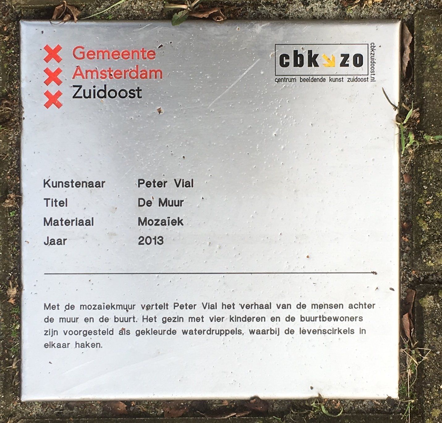 Witte kunstplaquette met Nederlandse tekst en een CBK-logo, liggend op gras met bladeren eromheen.