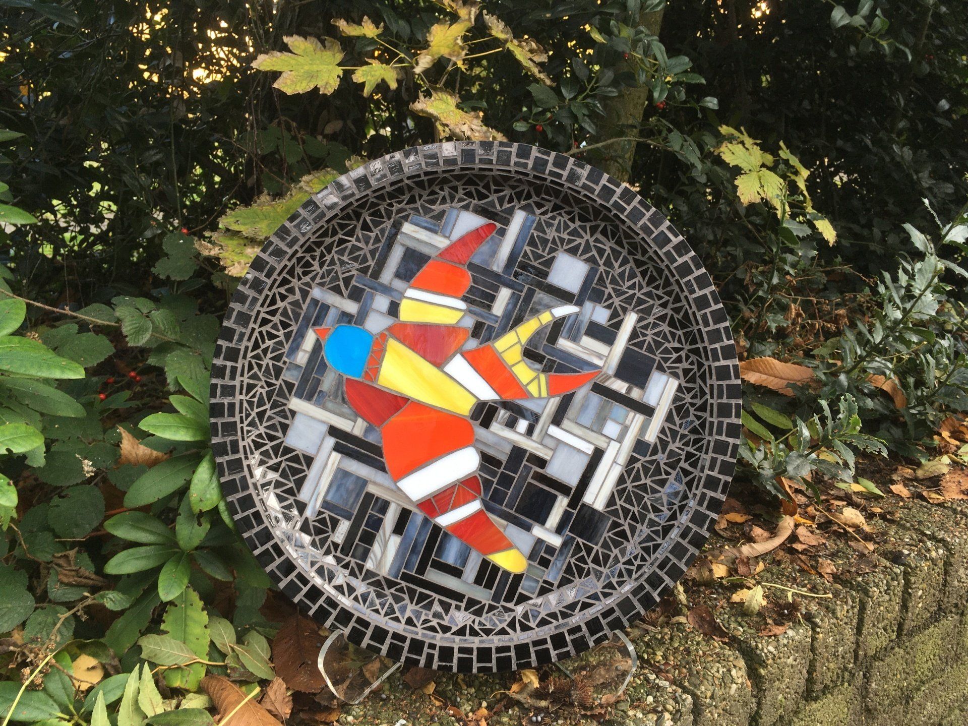 Rond mozaïekkunstwerk met een abstract patroon in rood, oranje en blauw, leunend tegen een tuinmuur.