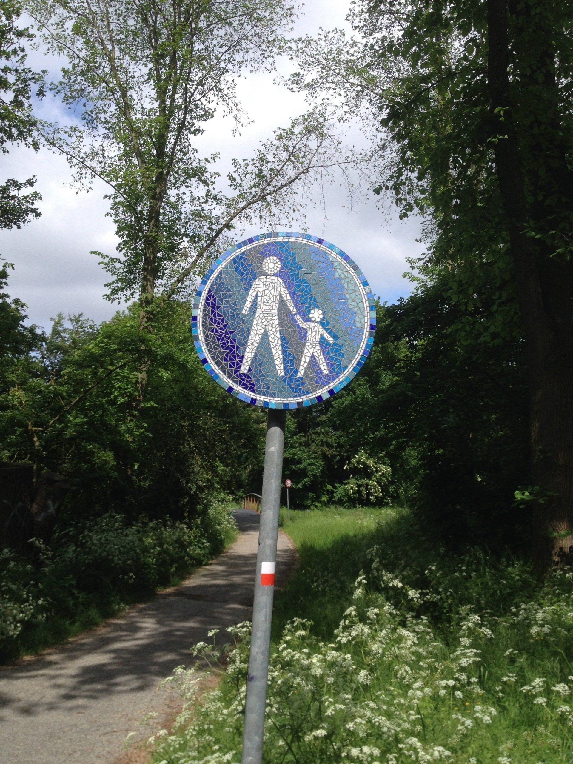 Blauw voetgangersbord op een pad in een lommerrijk park, omringd door wilde bloemen en bomen.