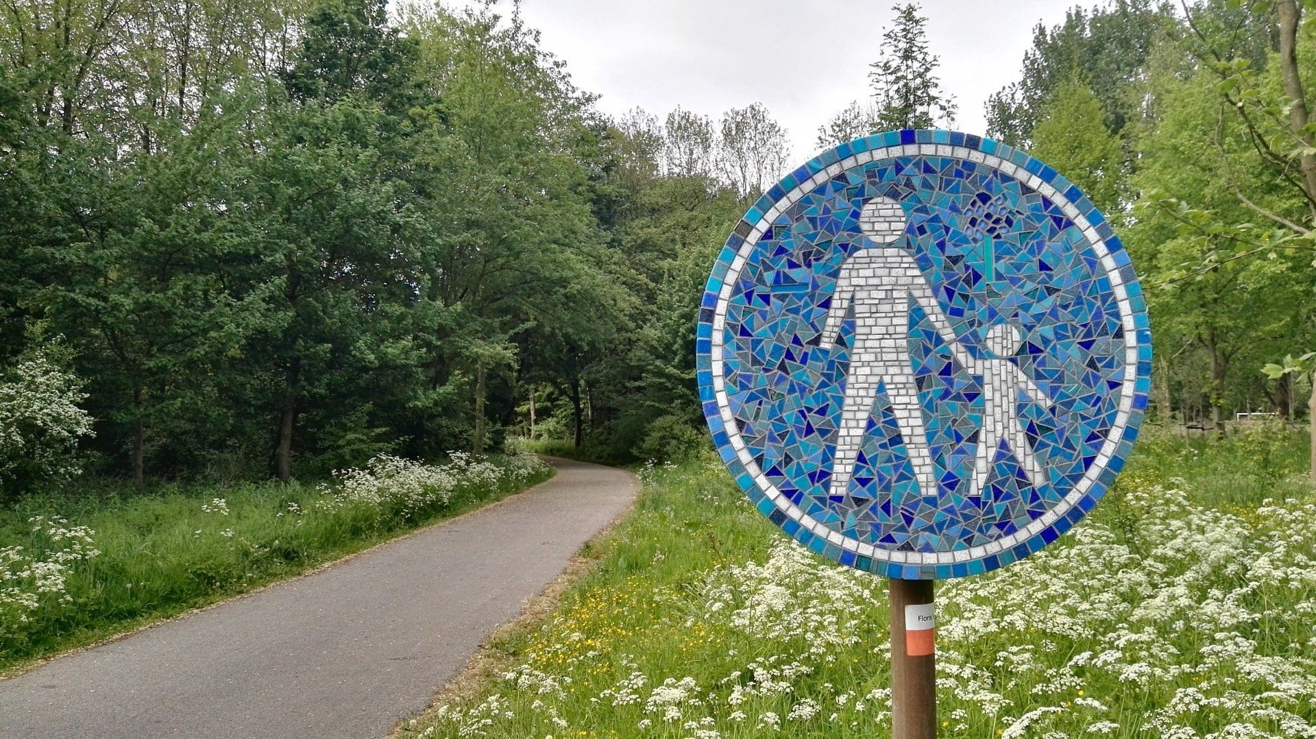 Blauw voetgangersoversteekbord naast een verhard pad in een groen park.