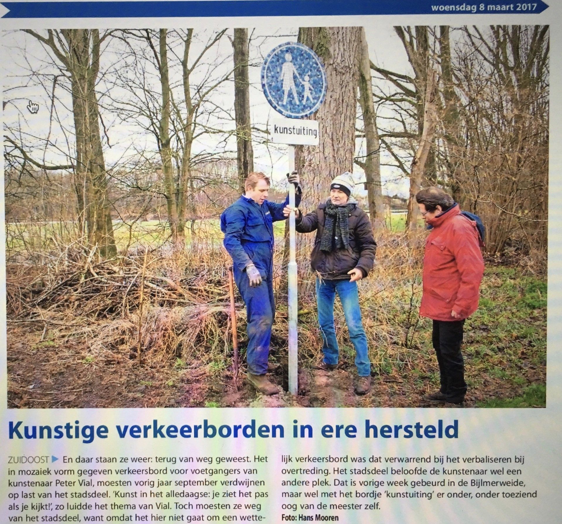Drie mensen staan bij een blauw voetgangersbord in een bosrijk gebied, waarvan er één omhoog wijst.