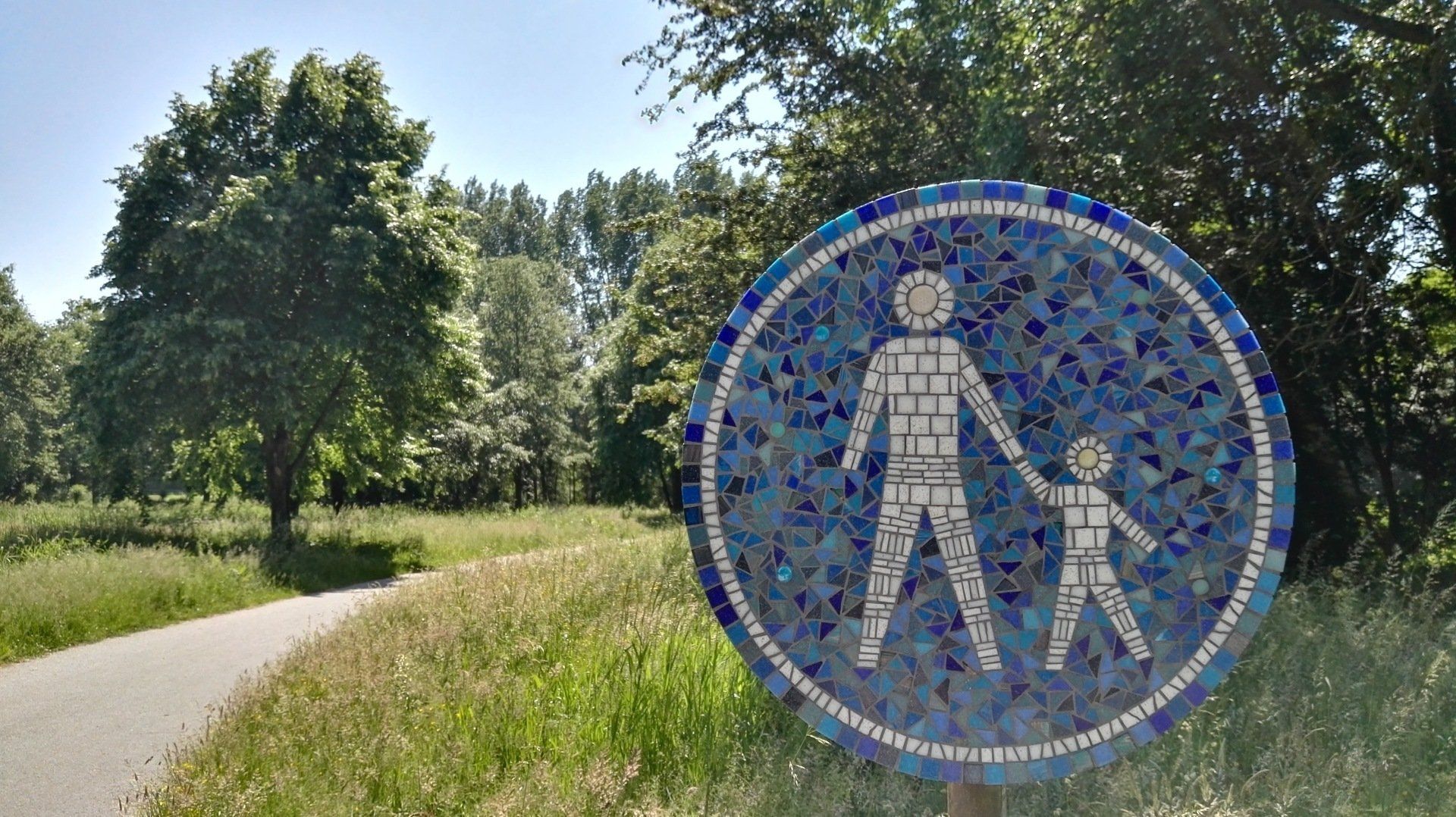 Blauw mozaïek voetgangersbord langs een pad in een zonnig groen park.