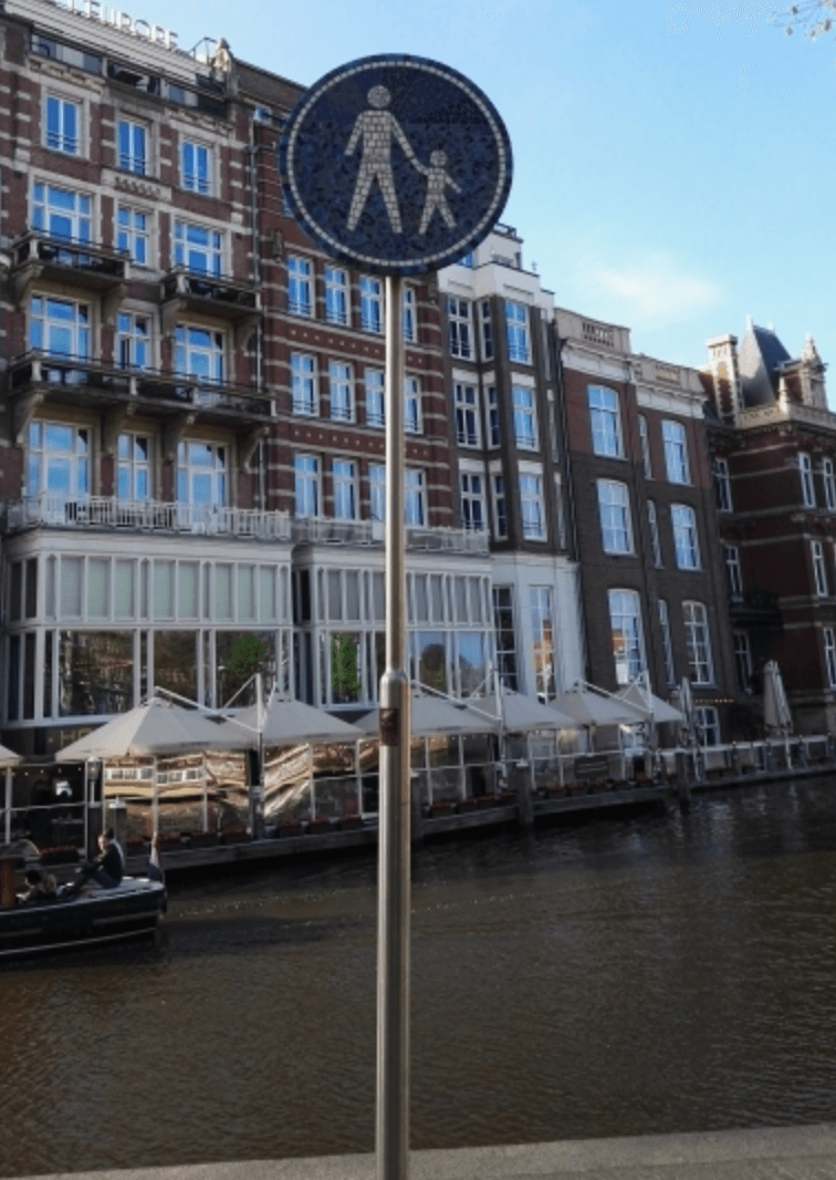 Voetgangersoversteekplaatsbord bij een kanaal met bakstenen gebouwen en aangemeerde boten op de achtergrond.