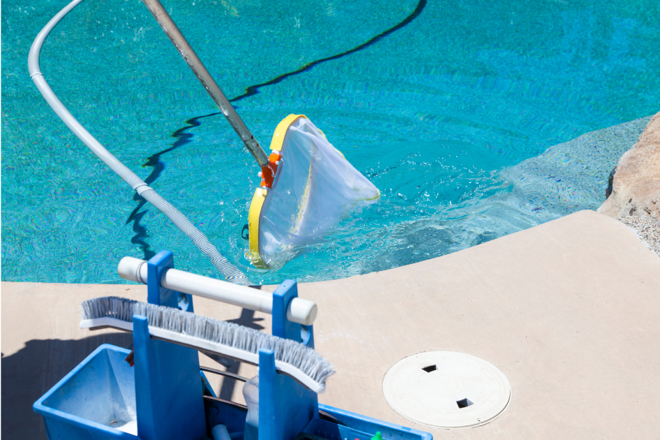 A person in blue overalls cleans a pool with a long-handled brush next to a house.