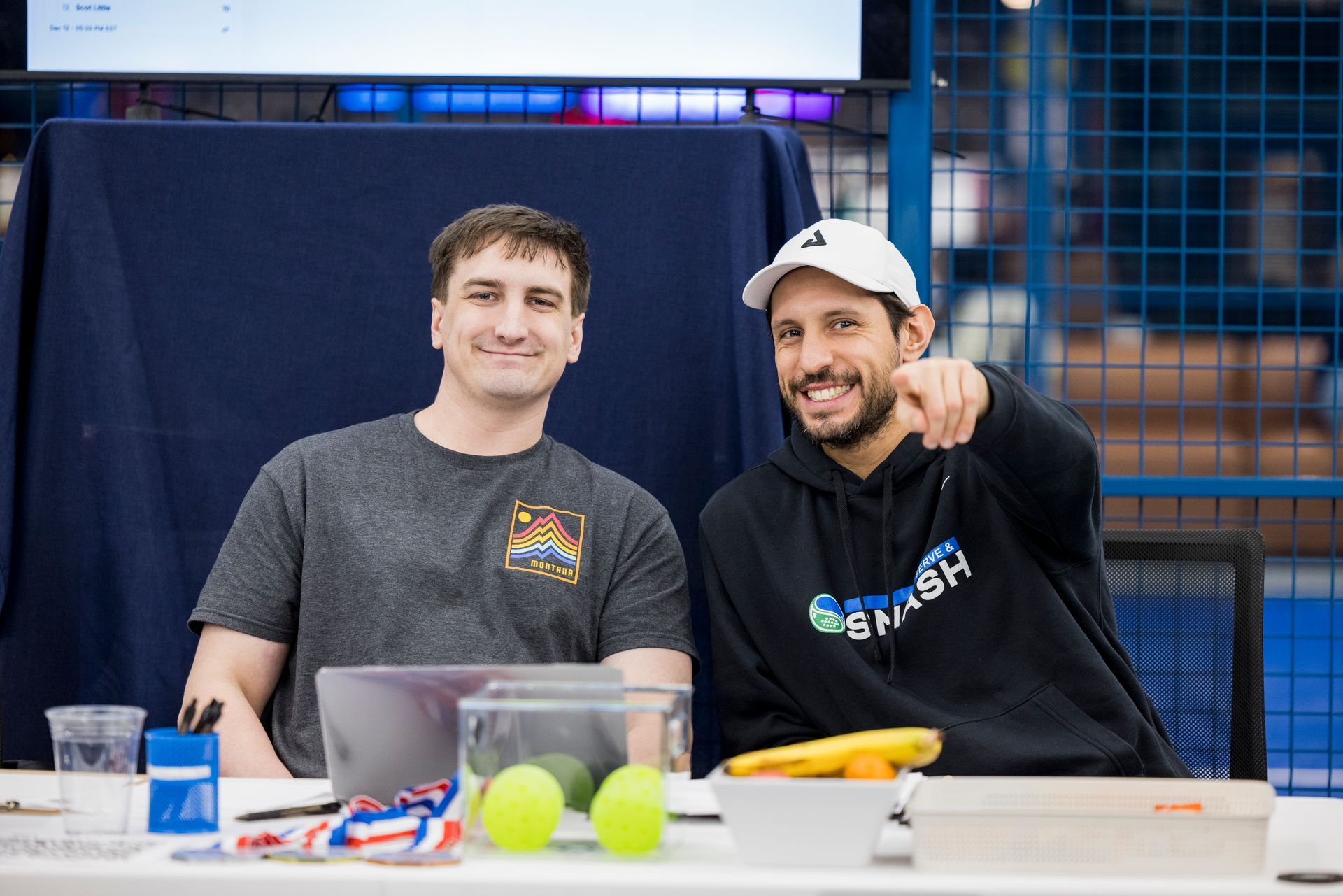 Two people sit at a table with a laptop and tennis balls; one points forward, while the other smiles at the camera.