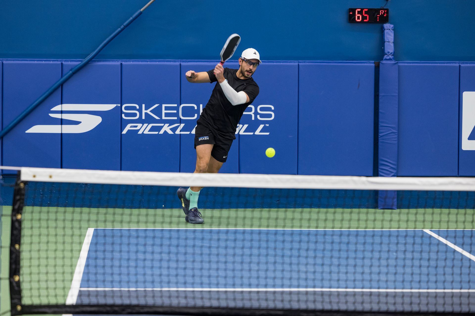A person in a black athletic outfit swings a pickleball paddle to hit a ball on an indoor blue court.