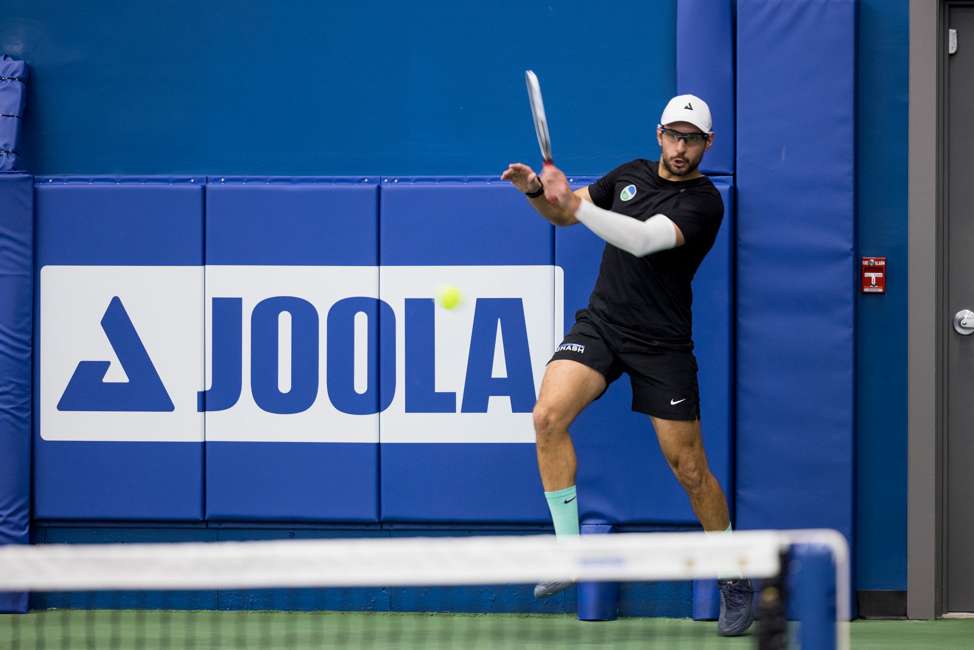A pickleball player in black athletic wear hitting a ball in front of a blue JOOLA-branded padded wall.