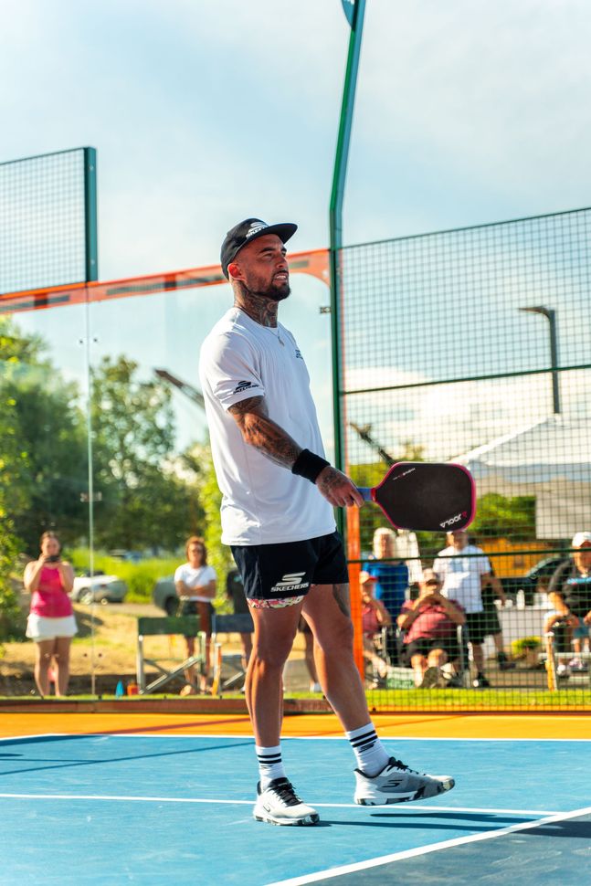 A person in a white t-shirt and black shorts holds a pickleball paddle on a blue outdoor court, looking toward the side.