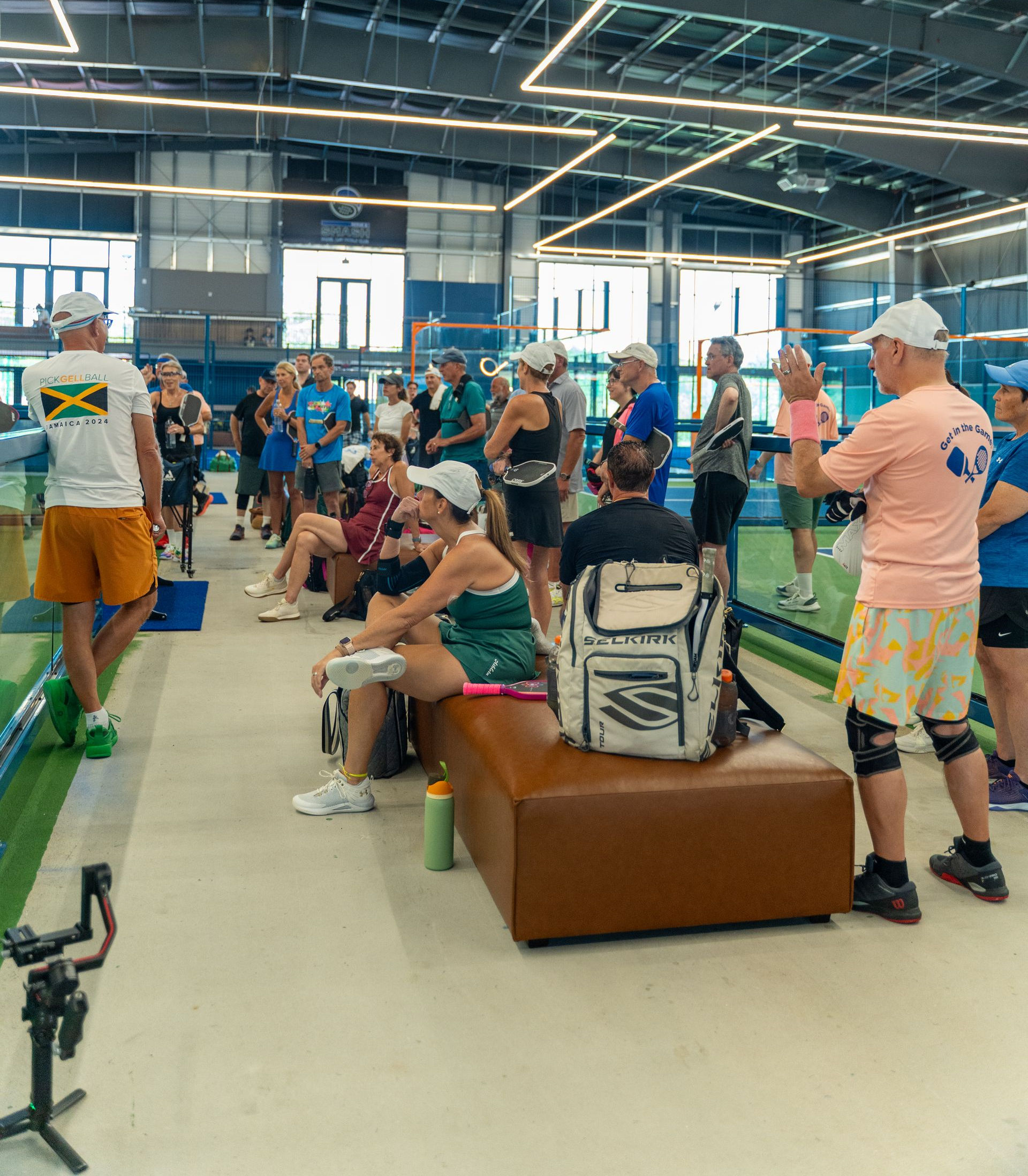 People gathered in an indoor pool area, some standing and chatting, with a seated person on a bench.