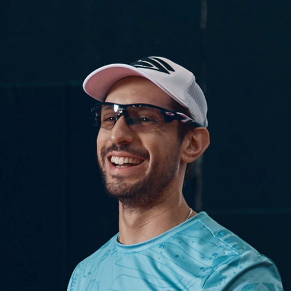 A smiling person wears a white baseball cap, dark sports sunglasses, and a light blue patterned athletic shirt.