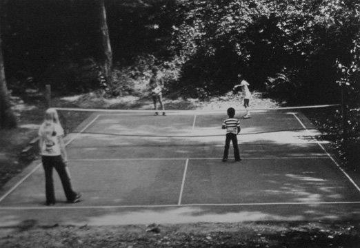 Four people play on a small outdoor court with a low net in a wooded, sunlit area.